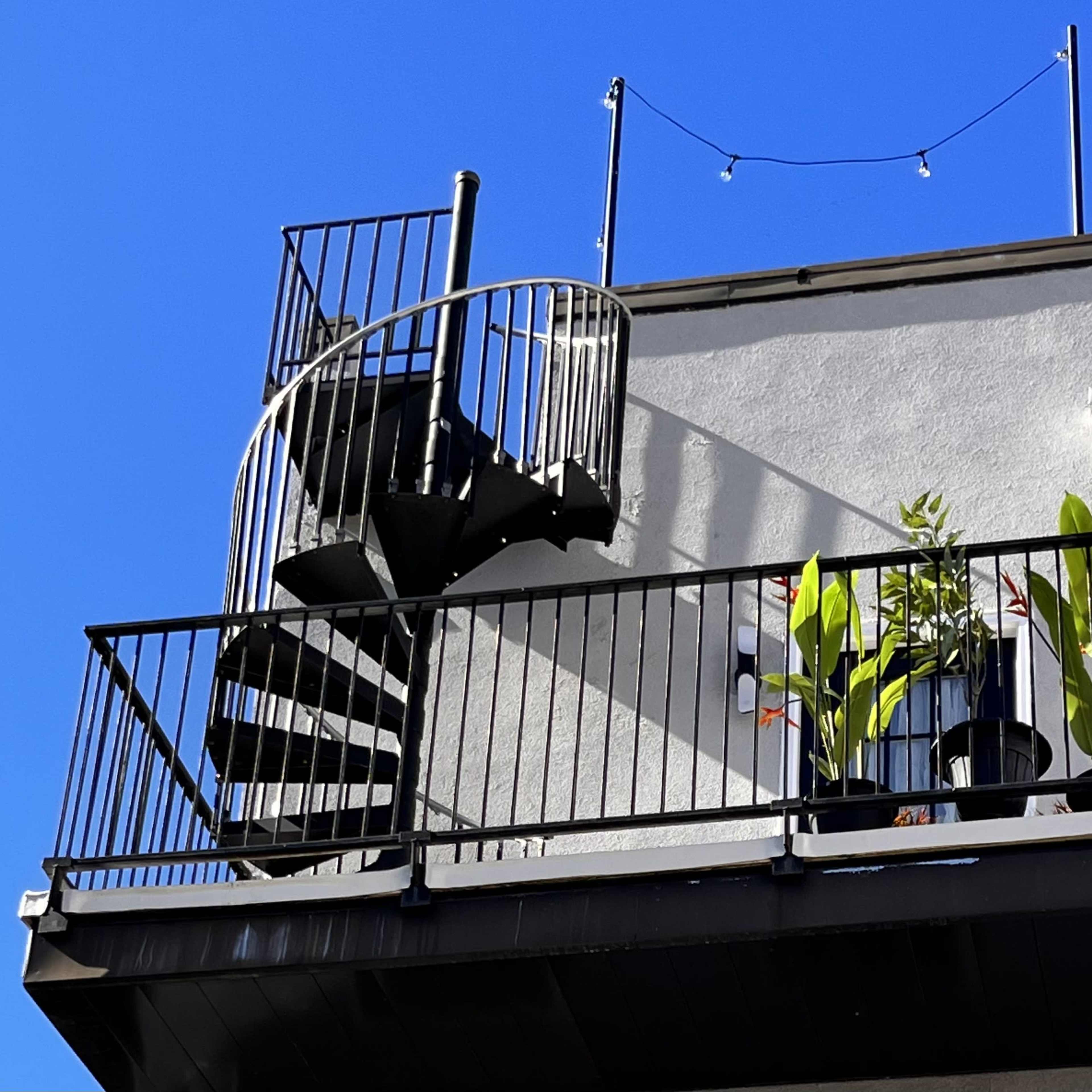A spiral staircase leads to a balcony with potted plants against a clear blue sky.
