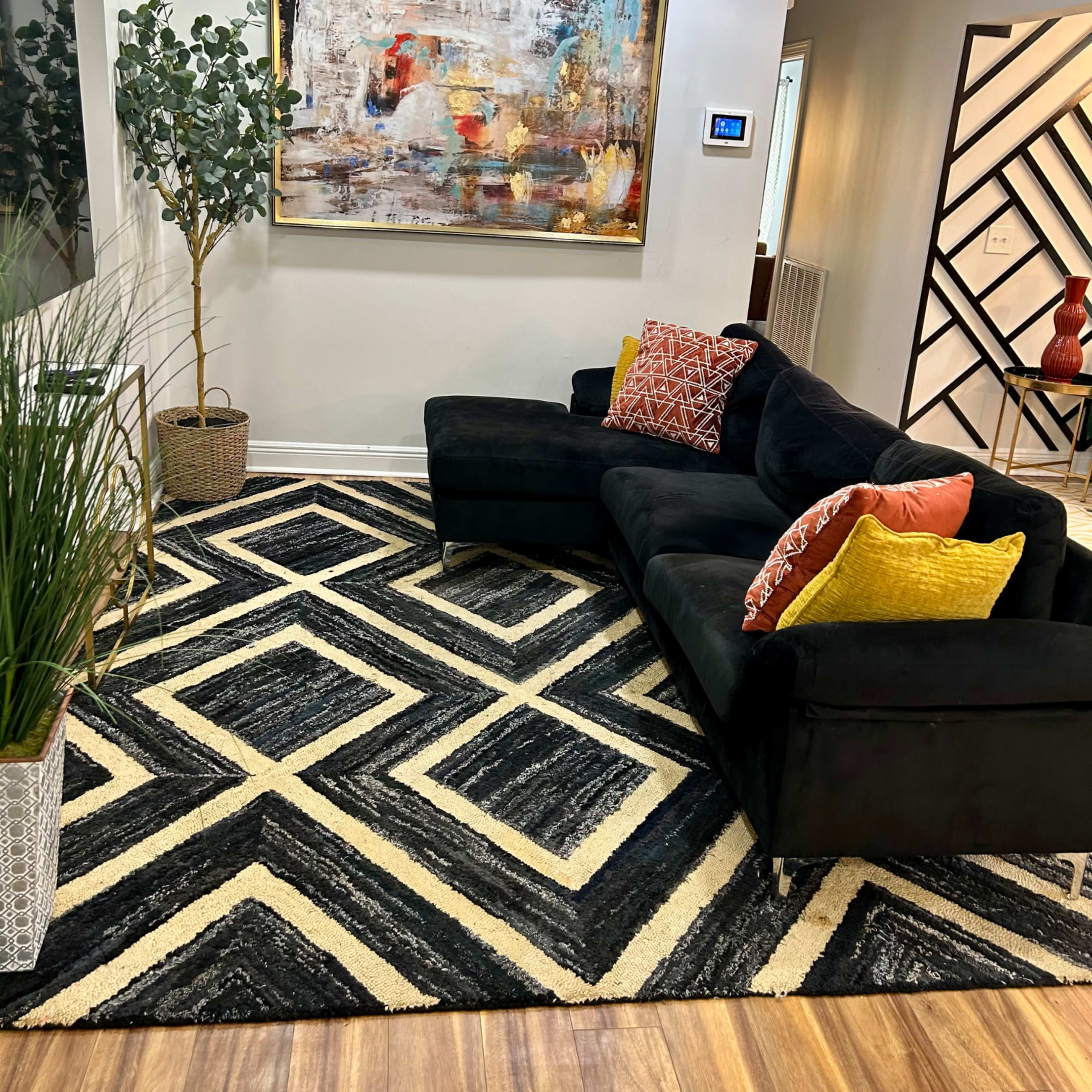A modern living room with a black sectional sofa, colorful cushions, a large abstract painting on the wall, a patterned rug, and decorative plants.