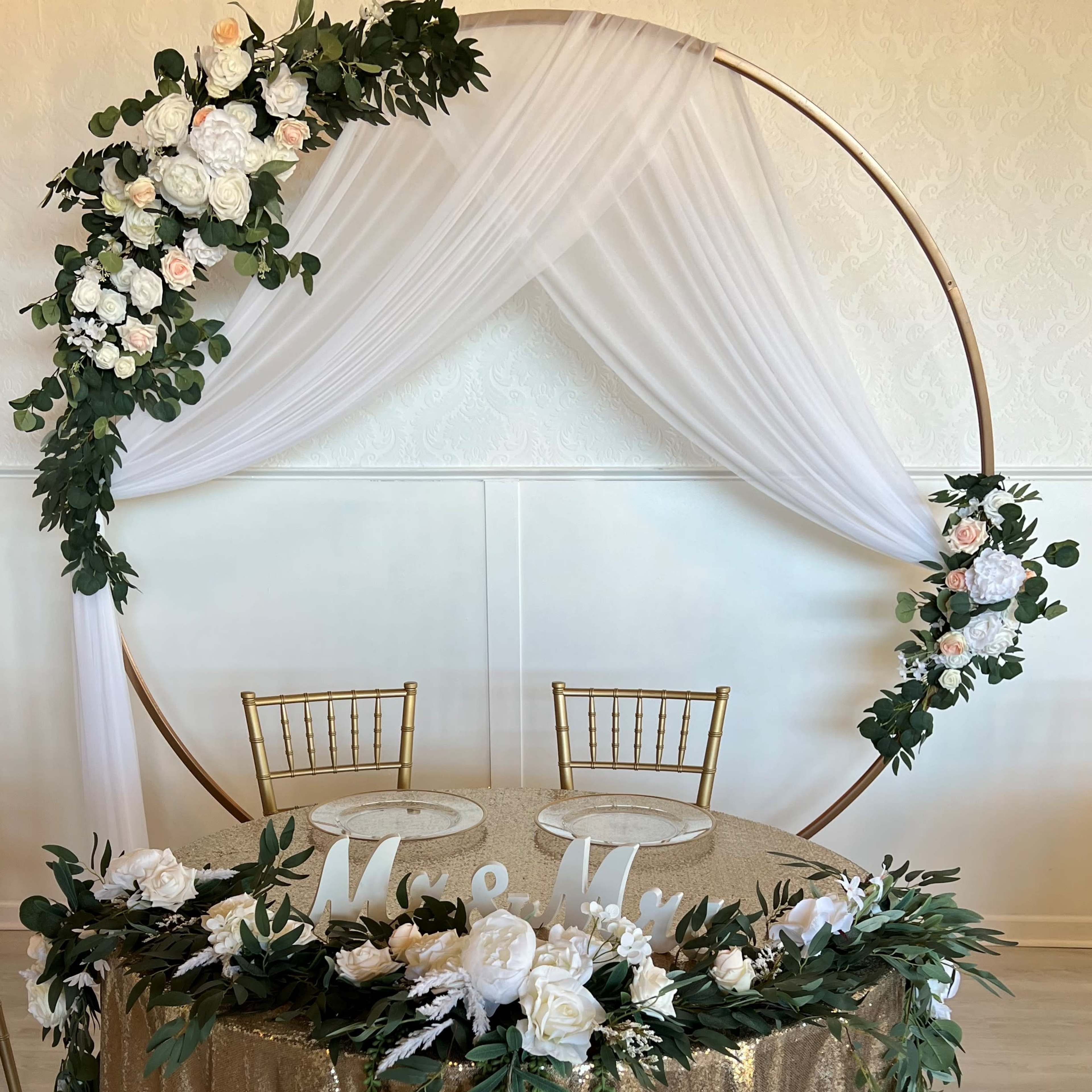 A round floral arch is decorated with white and pink flowers, framing a table with a gold sequin cloth and two empty gold chairs.