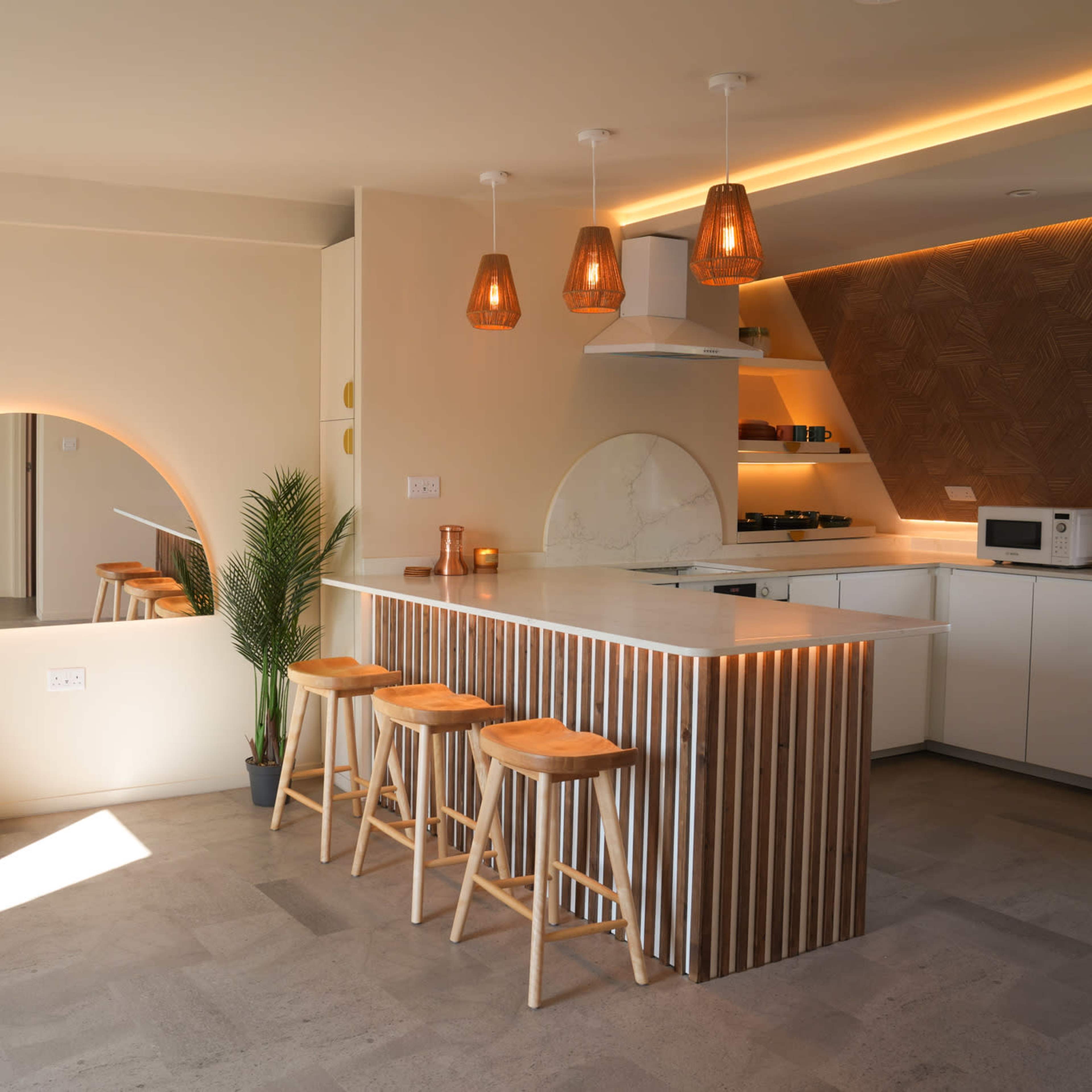 A modern kitchen with a central island made of slatted wood, three wooden stools, and pendant lighting, complemented by a curved archway and a decorative plant.
