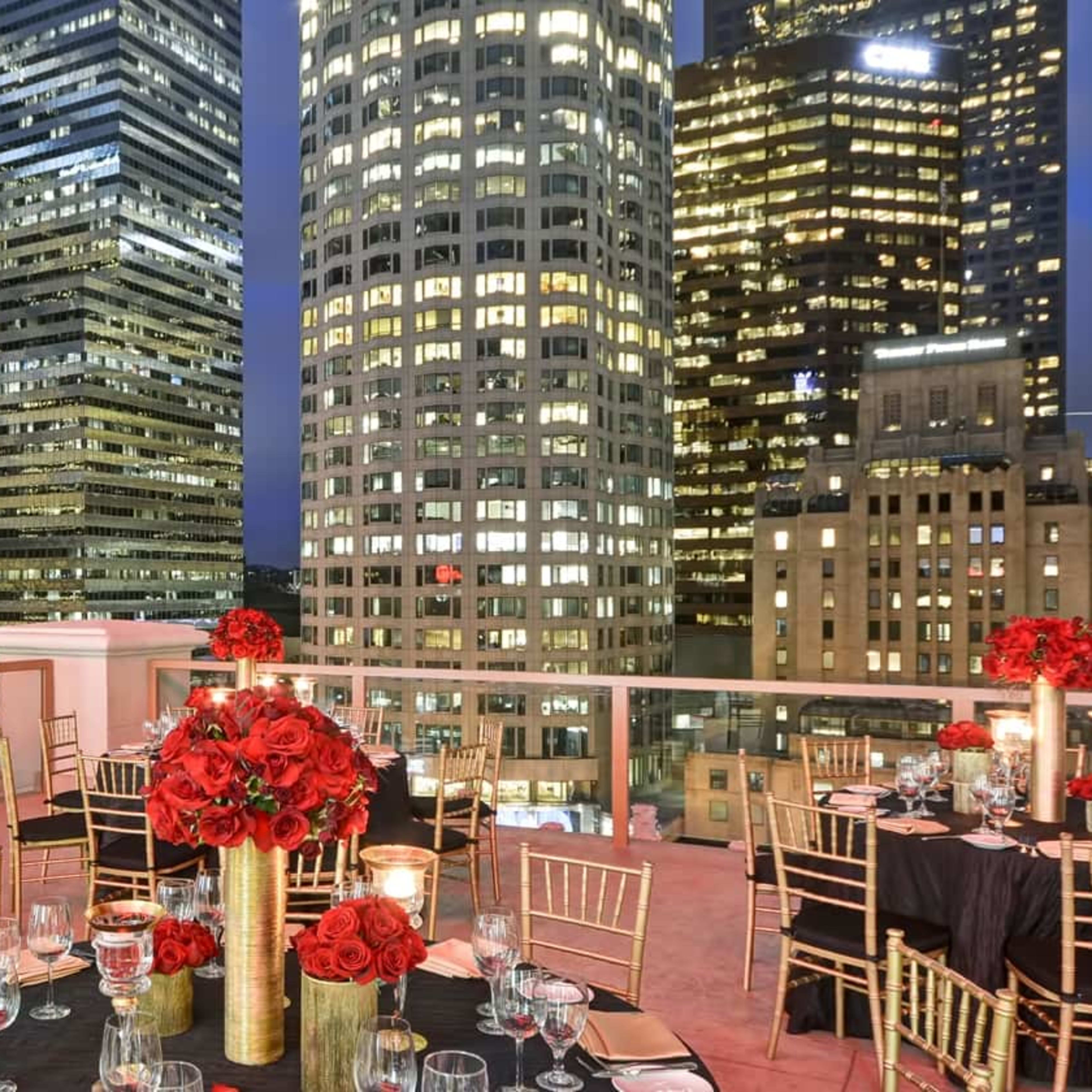A rooftop event space features elegantly set tables with red floral centerpieces and views of a city skyline illuminated at night.