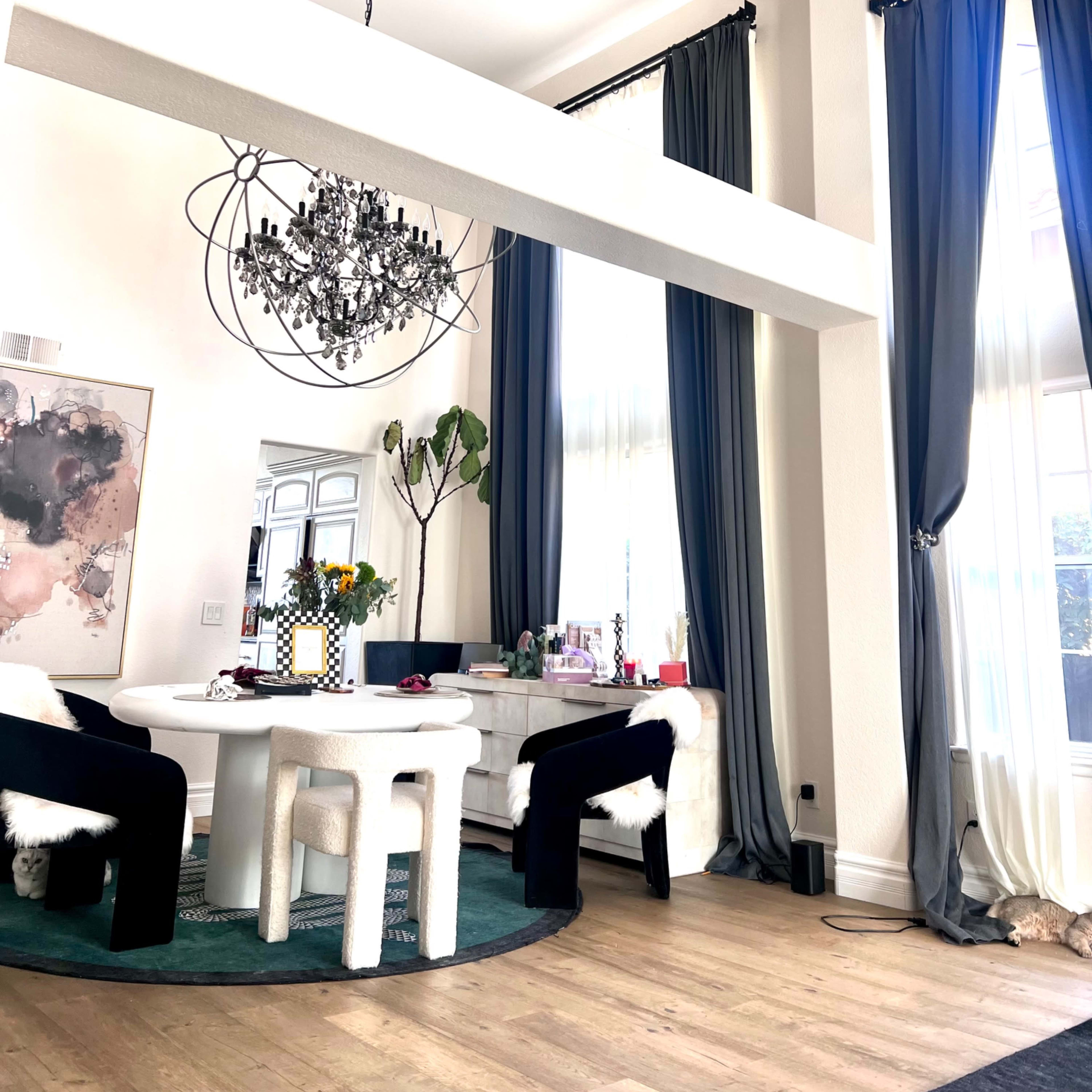 A modern dining area features a round table with black chairs, a decorative chandelier overhead, and large windows draped with blue and gray curtains.