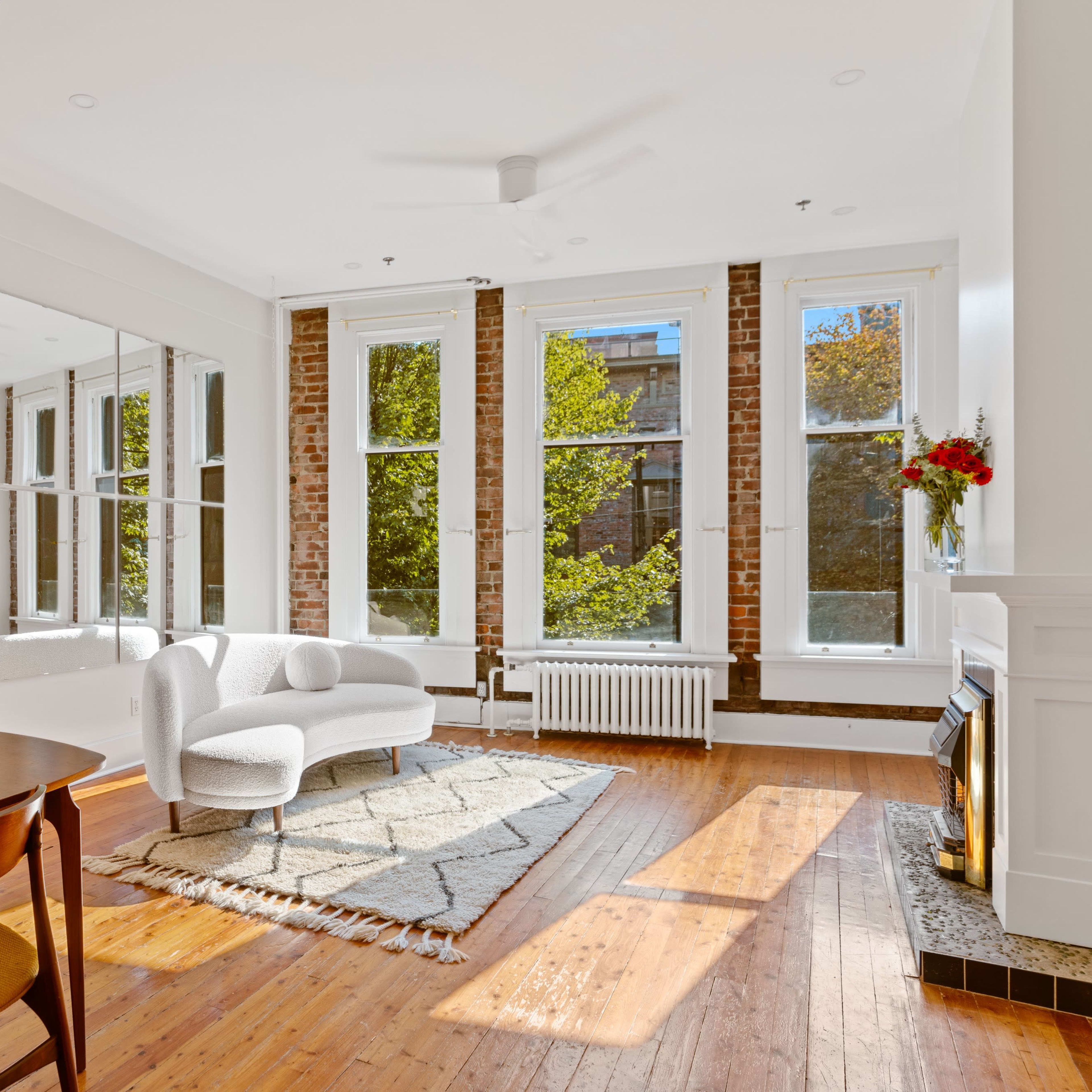 A bright living space features large windows, a sofa, and a dining table against a backdrop of exposed brick and a fireplace.
