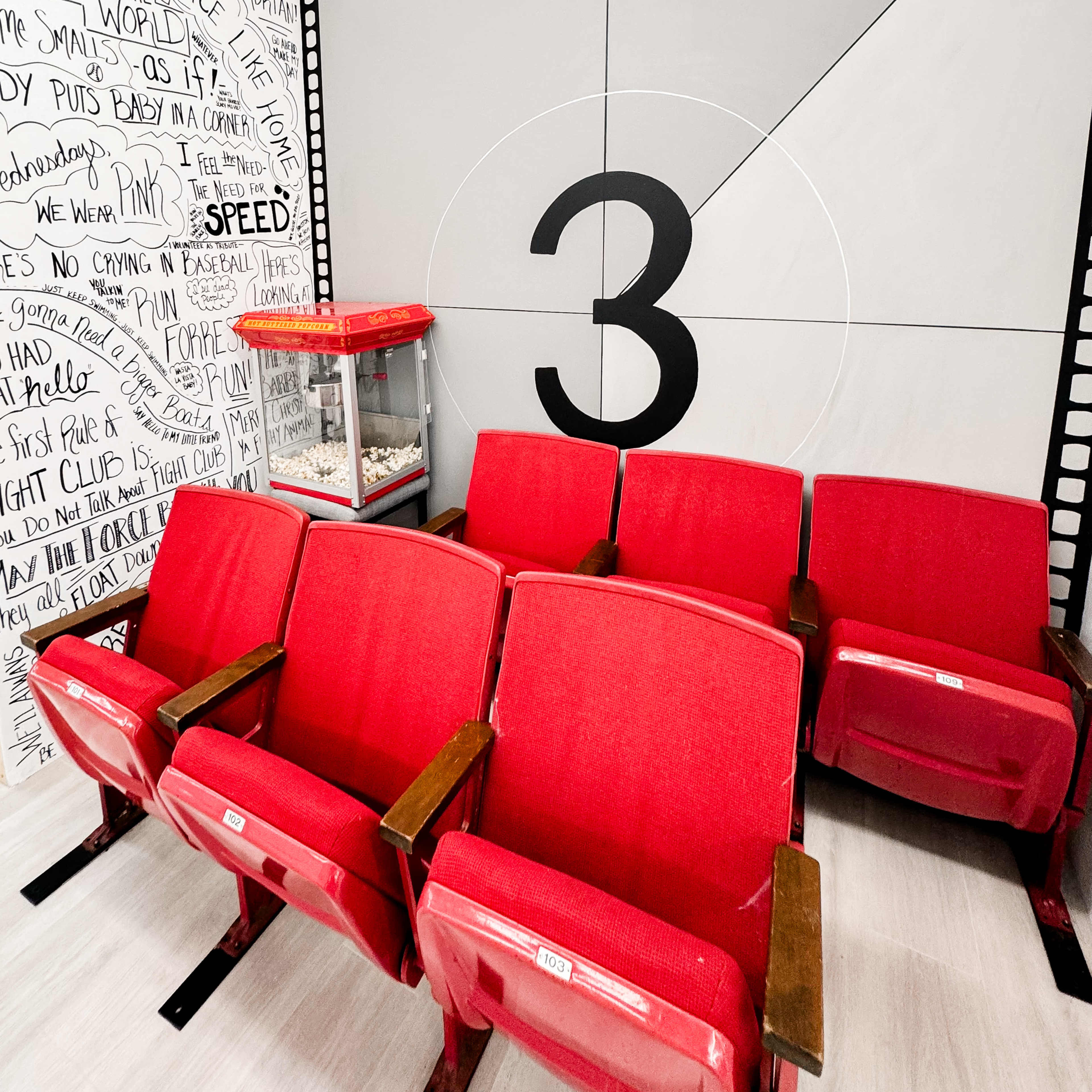 A row of red vintage theater seats in front of a large wall featuring a gray film slate with the number three and a backdrop of handwritten movie-themed phrases.