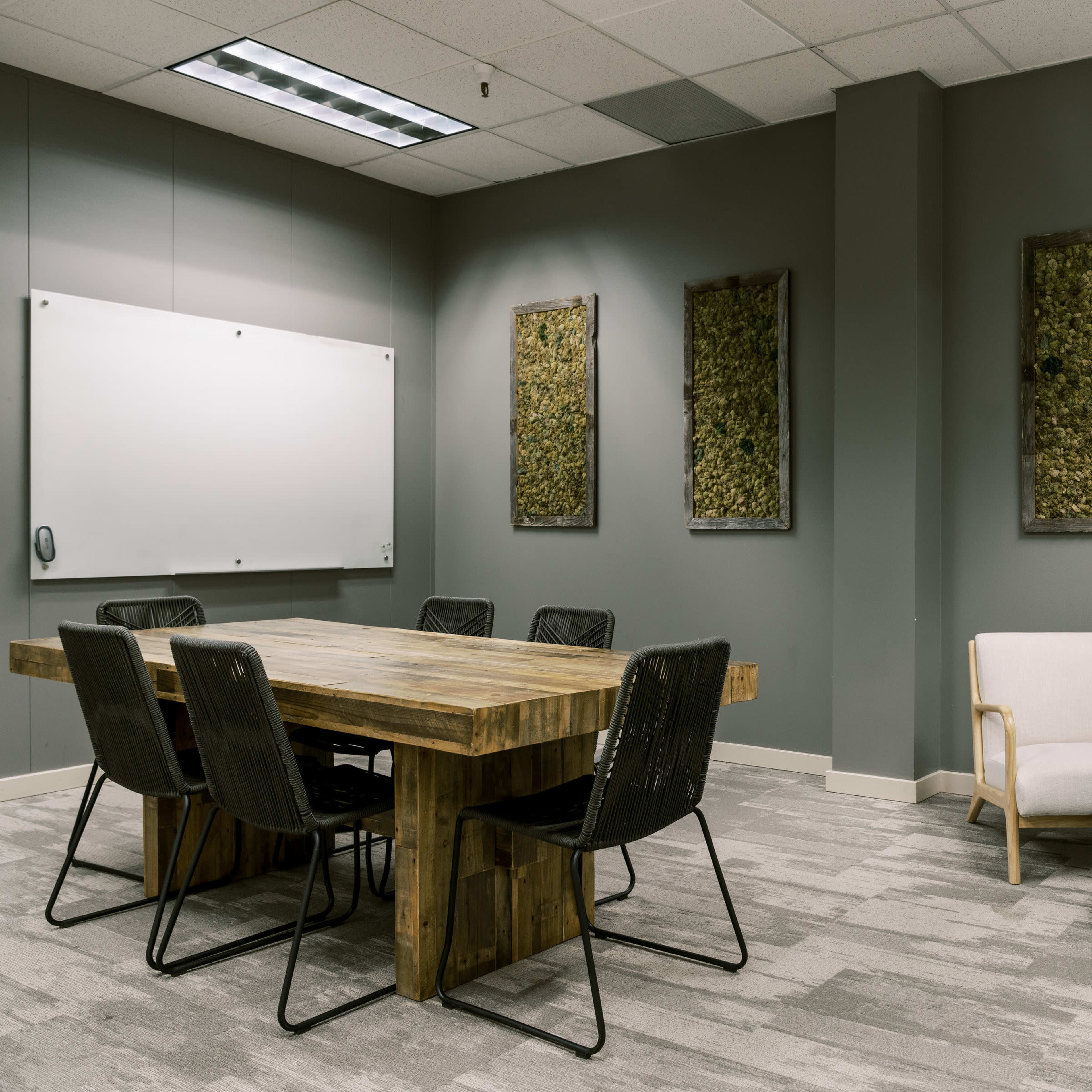 A conference room features a large wooden table surrounded by black mesh chairs, with two light-colored armchairs and decorative wall art made of natural materials.