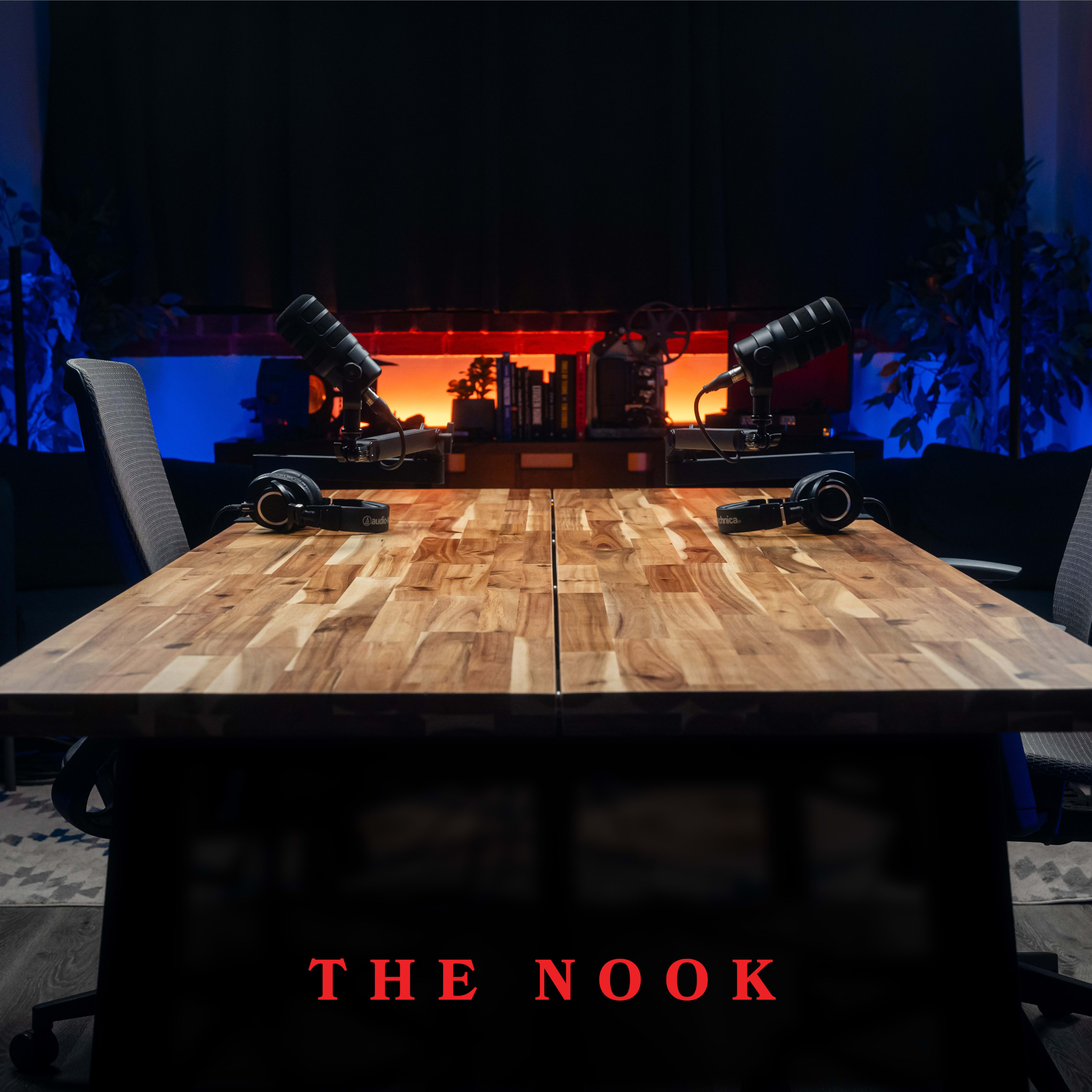 A wooden table with two chairs and microphones is set up for a podcast in a dimly lit room.