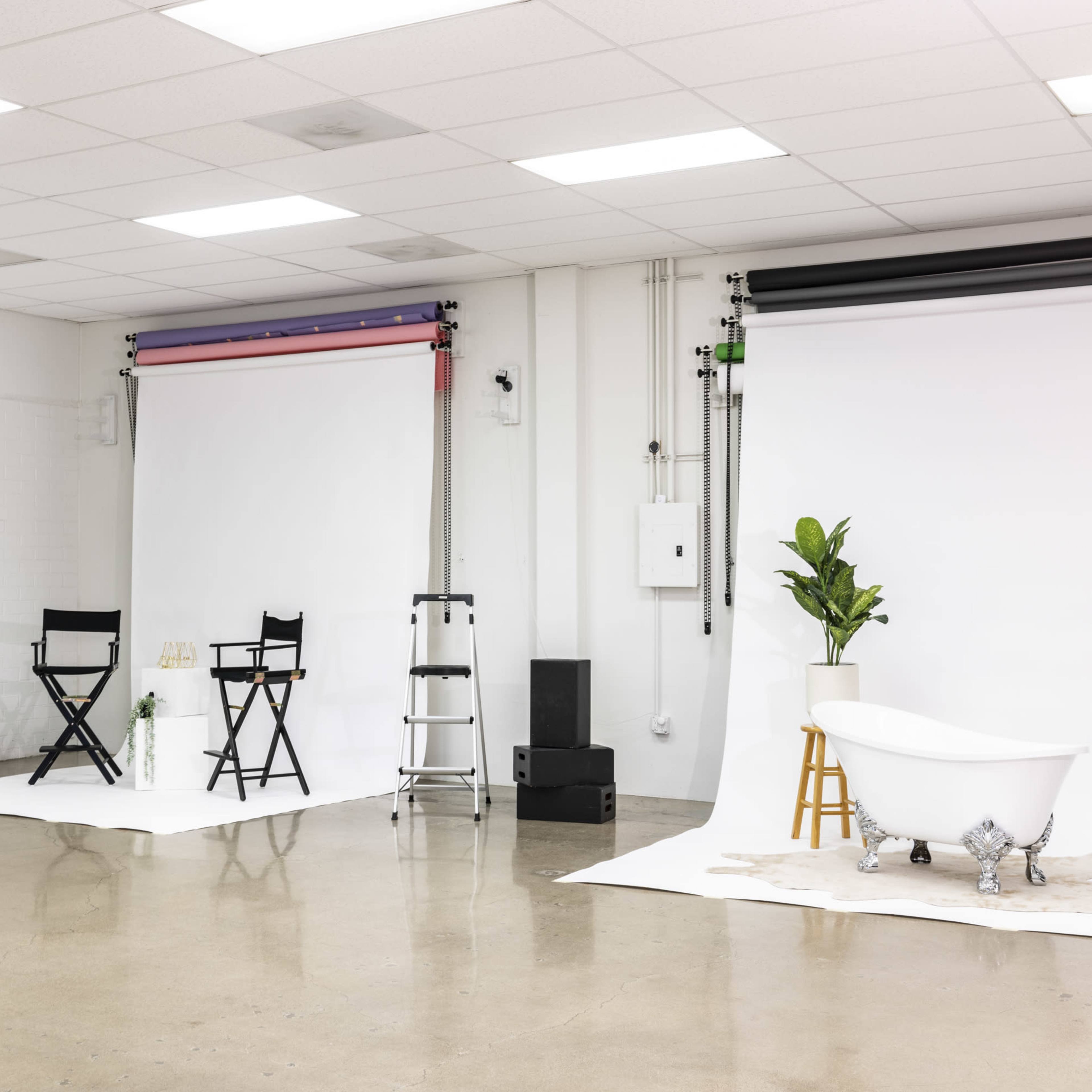 A photography studio with two backdrop rolls, a bathtub on a white backdrop, a potted plant, and various equipment including a black umbrella and director's chairs.
