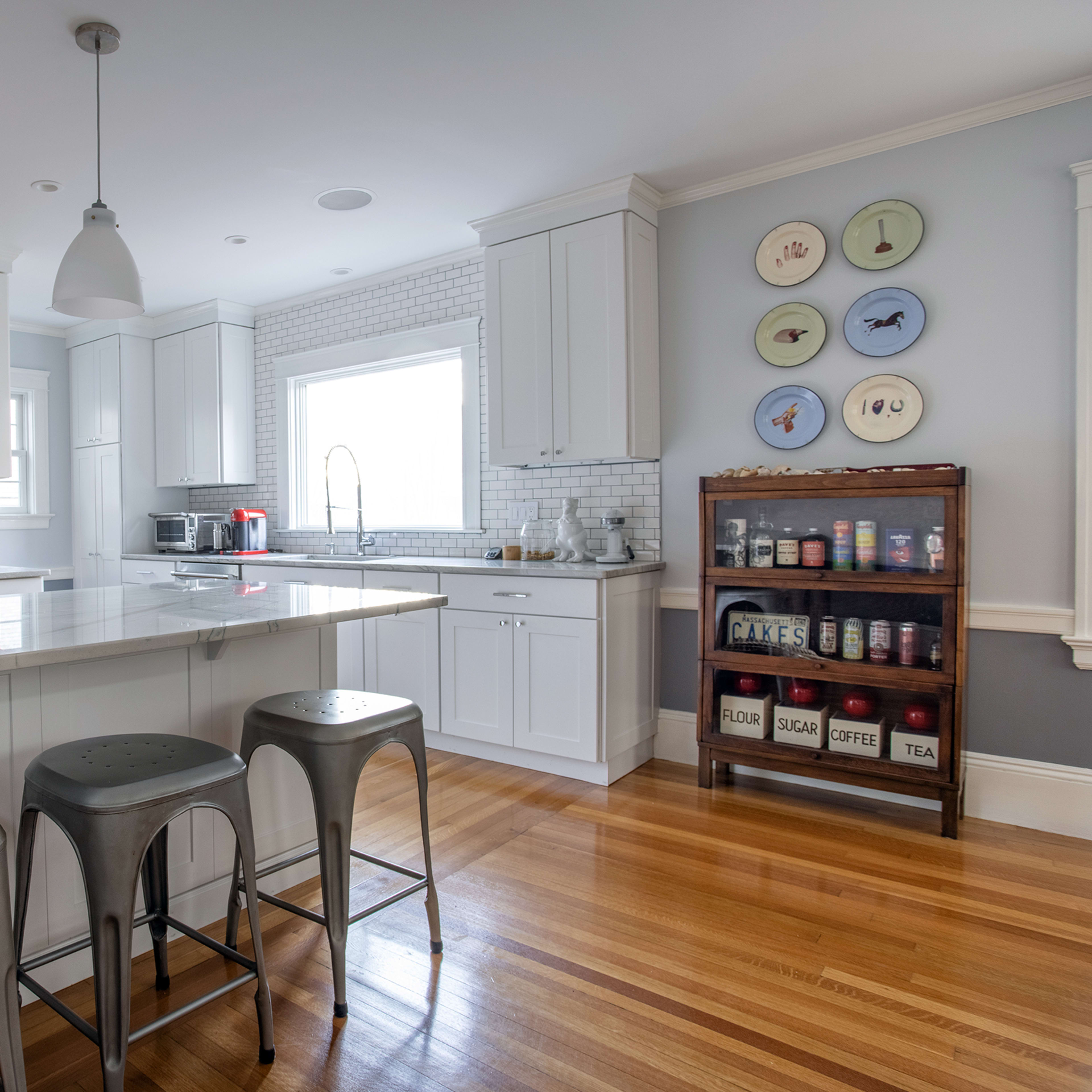 The kitchen features white cabinetry, a marble countertop island with three metal stools, and a wall of backsplash tiles.
