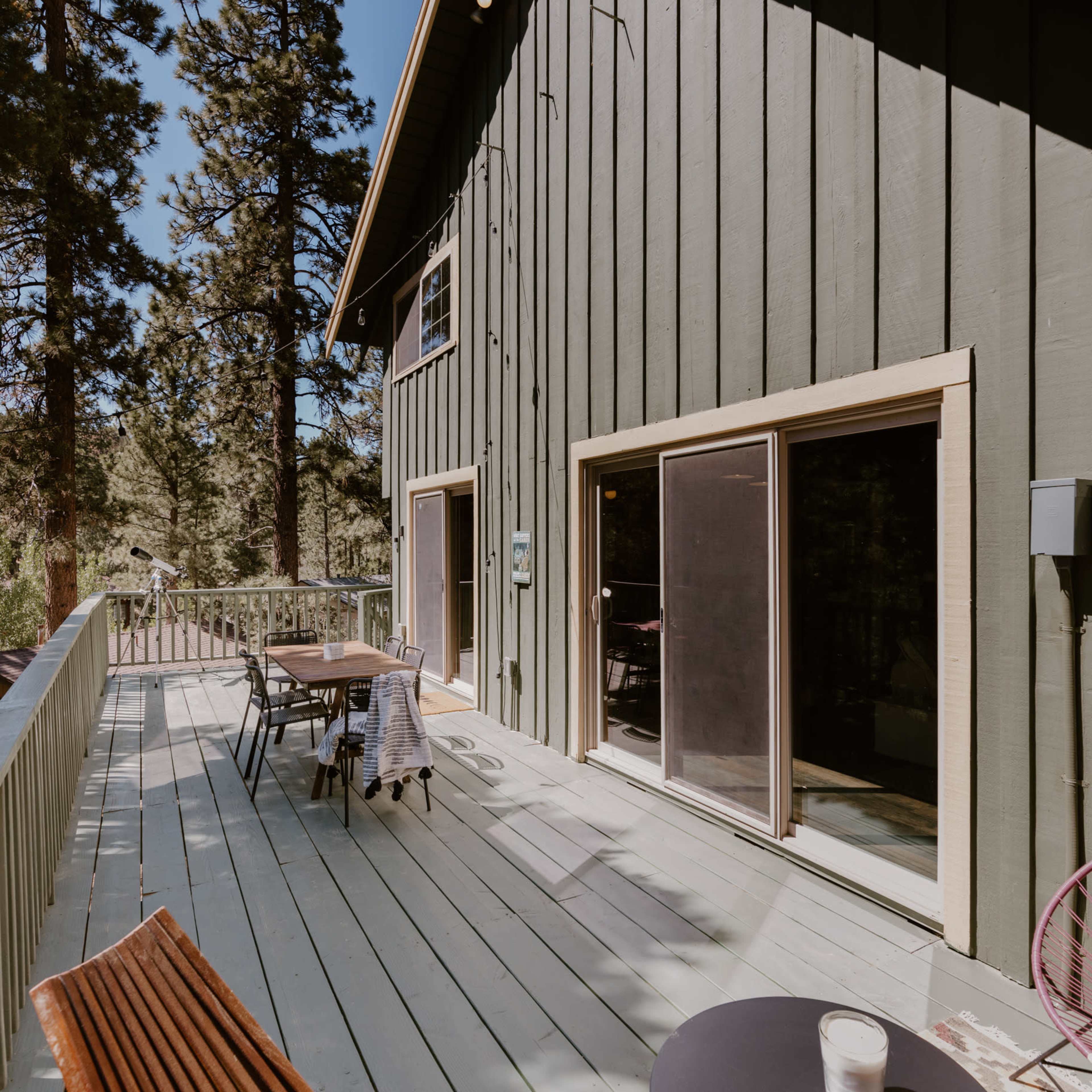 A green wooden cabin with a deck overlooks a forested area, featuring outdoor furniture and a screen door.