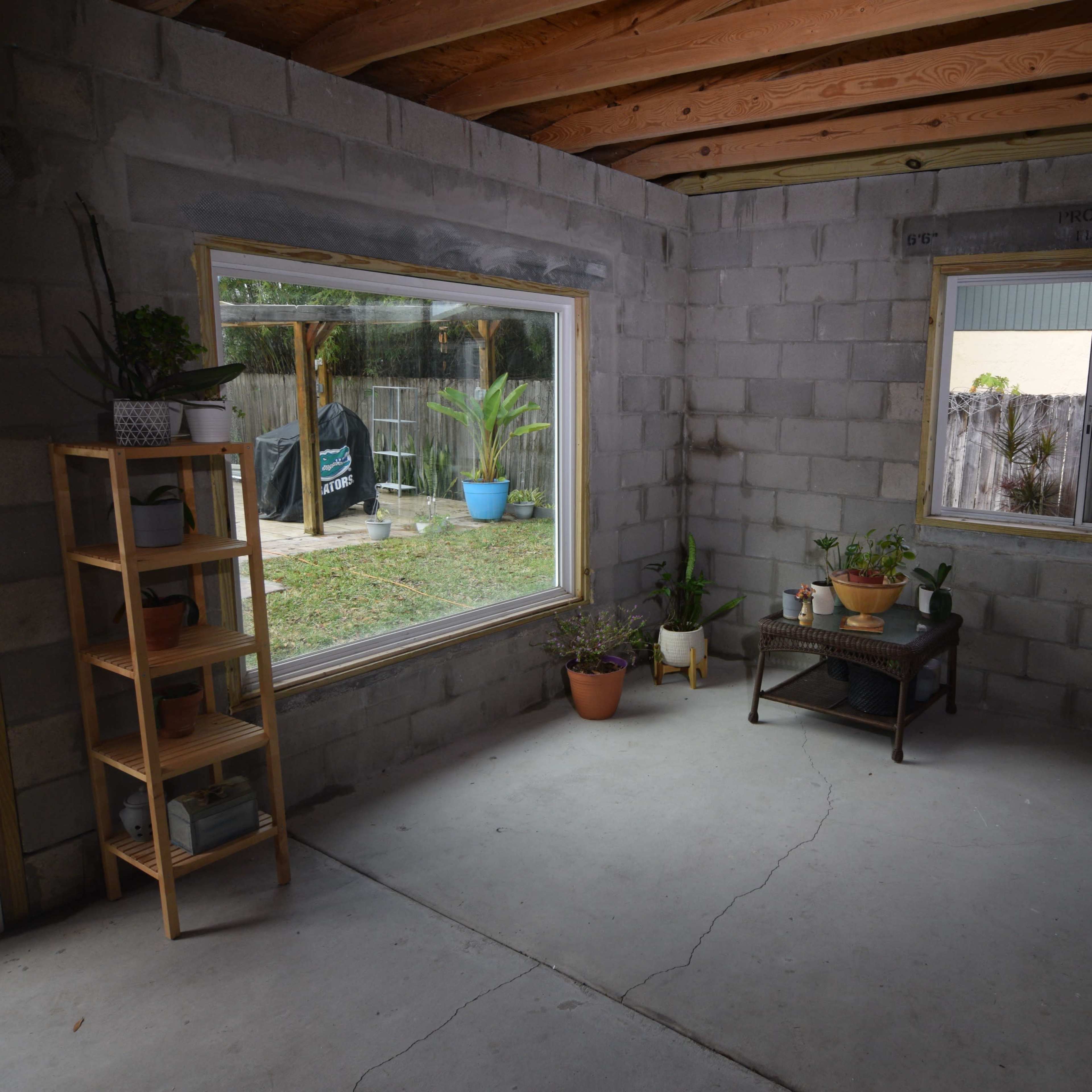 A concrete-walled room with two windows, a wooden shelf with potted plants, and a small table in the center.