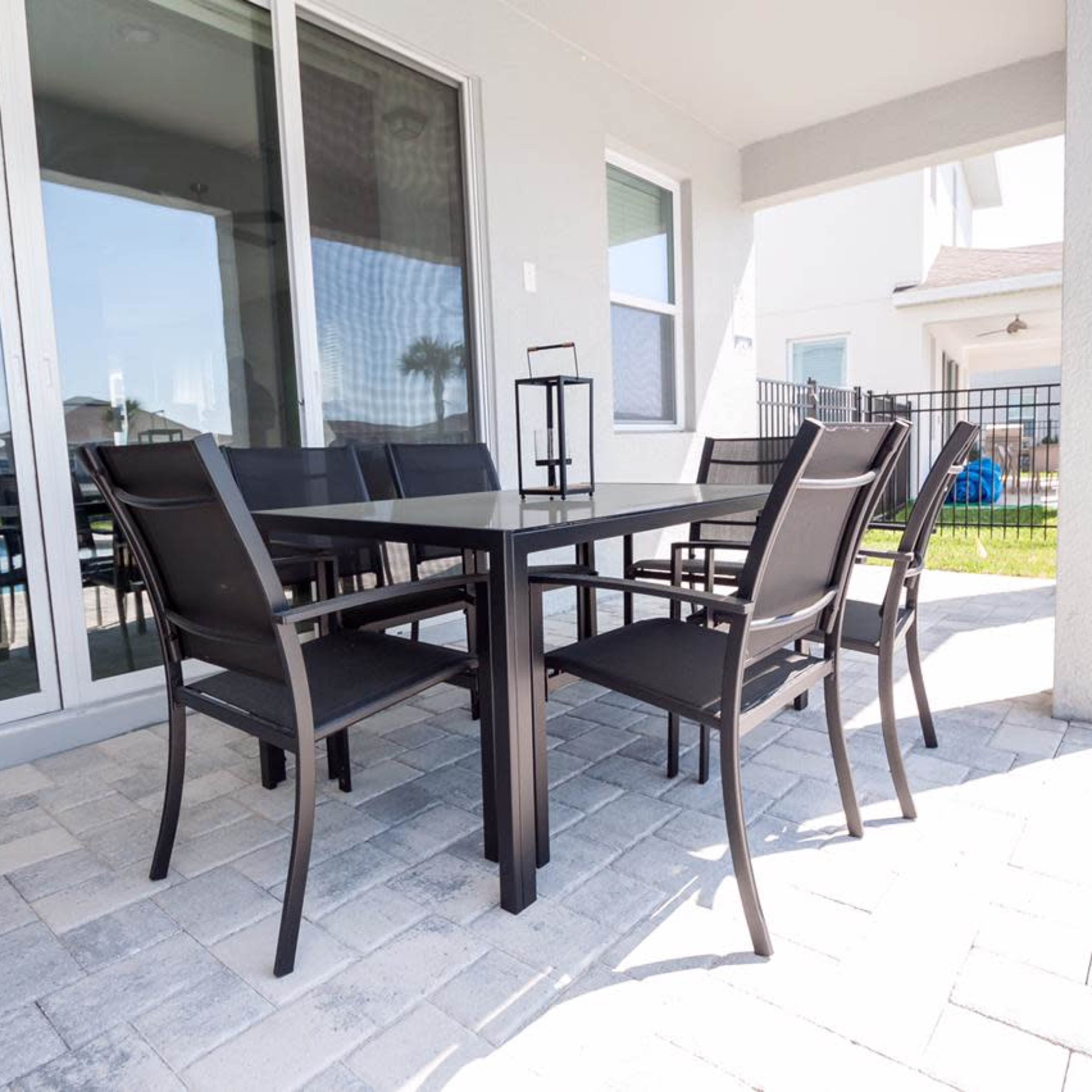 A patio area features a rectangular table surrounded by six chairs, with sliding glass doors in the background and a pool visible beyond a fence.