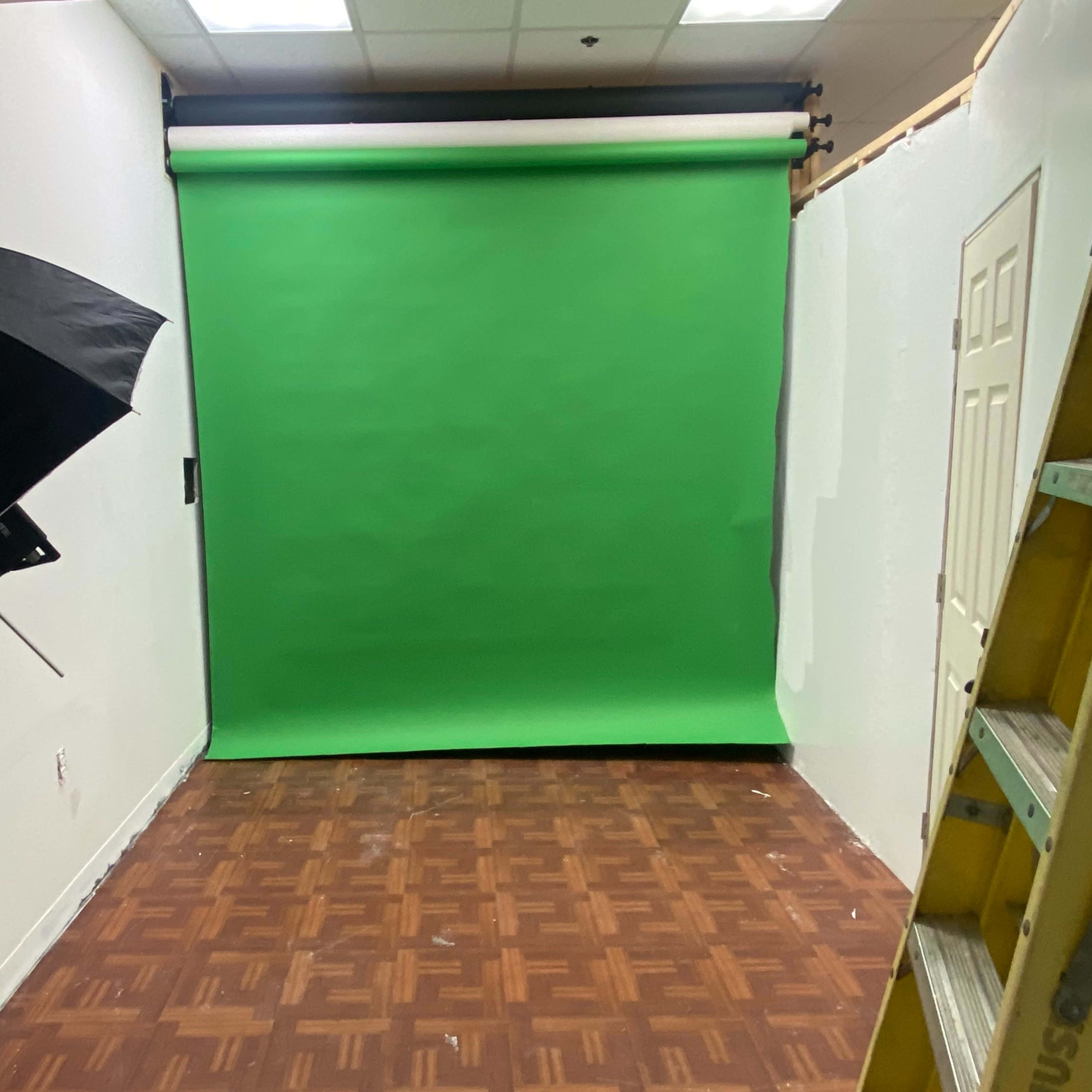 A small studio space with a green backdrop, a black umbrella light, and a ladder against the wall.