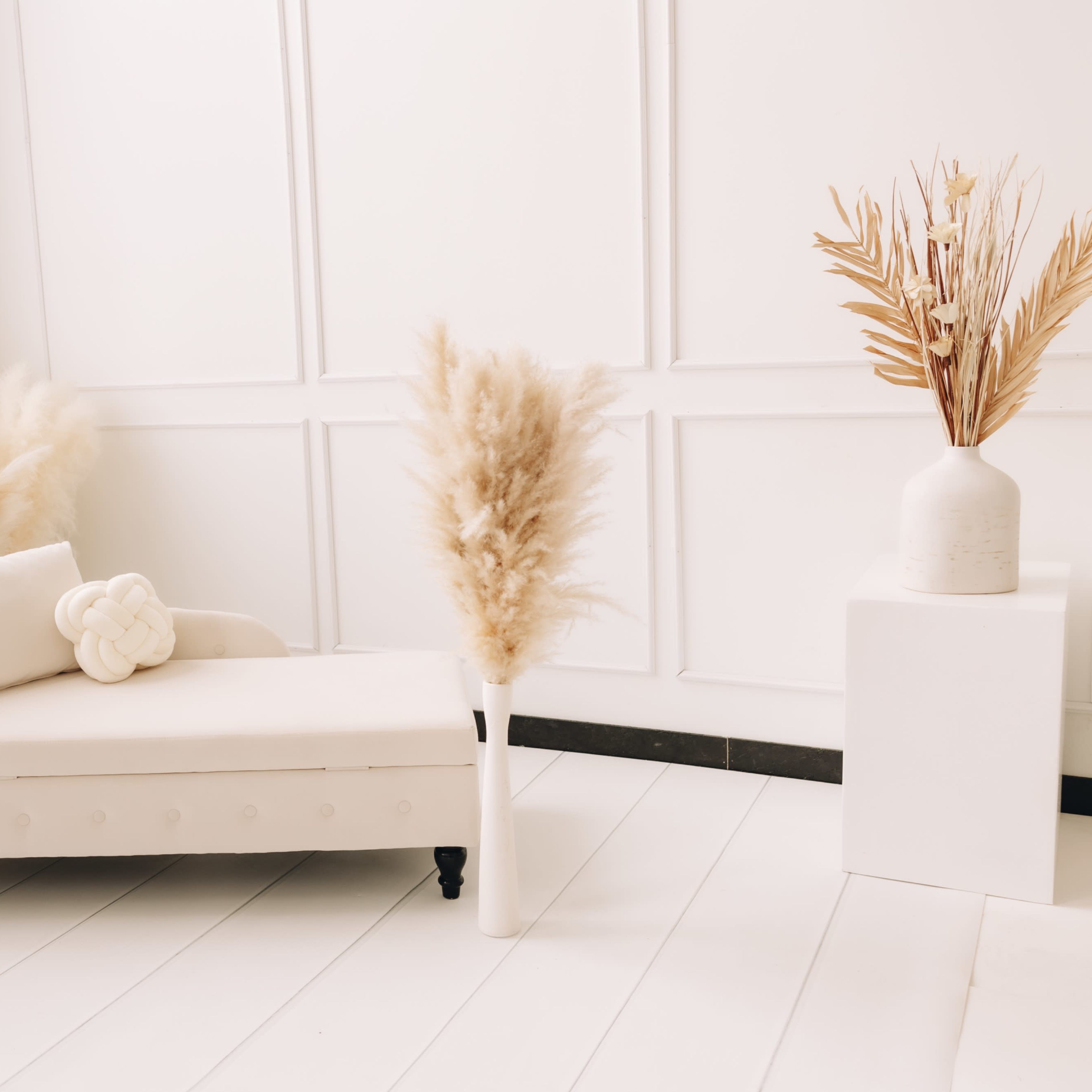 A minimalist, light-colored room featuring a chaise lounge, decorative pillows, and tall pampas grass arrangements in vases.