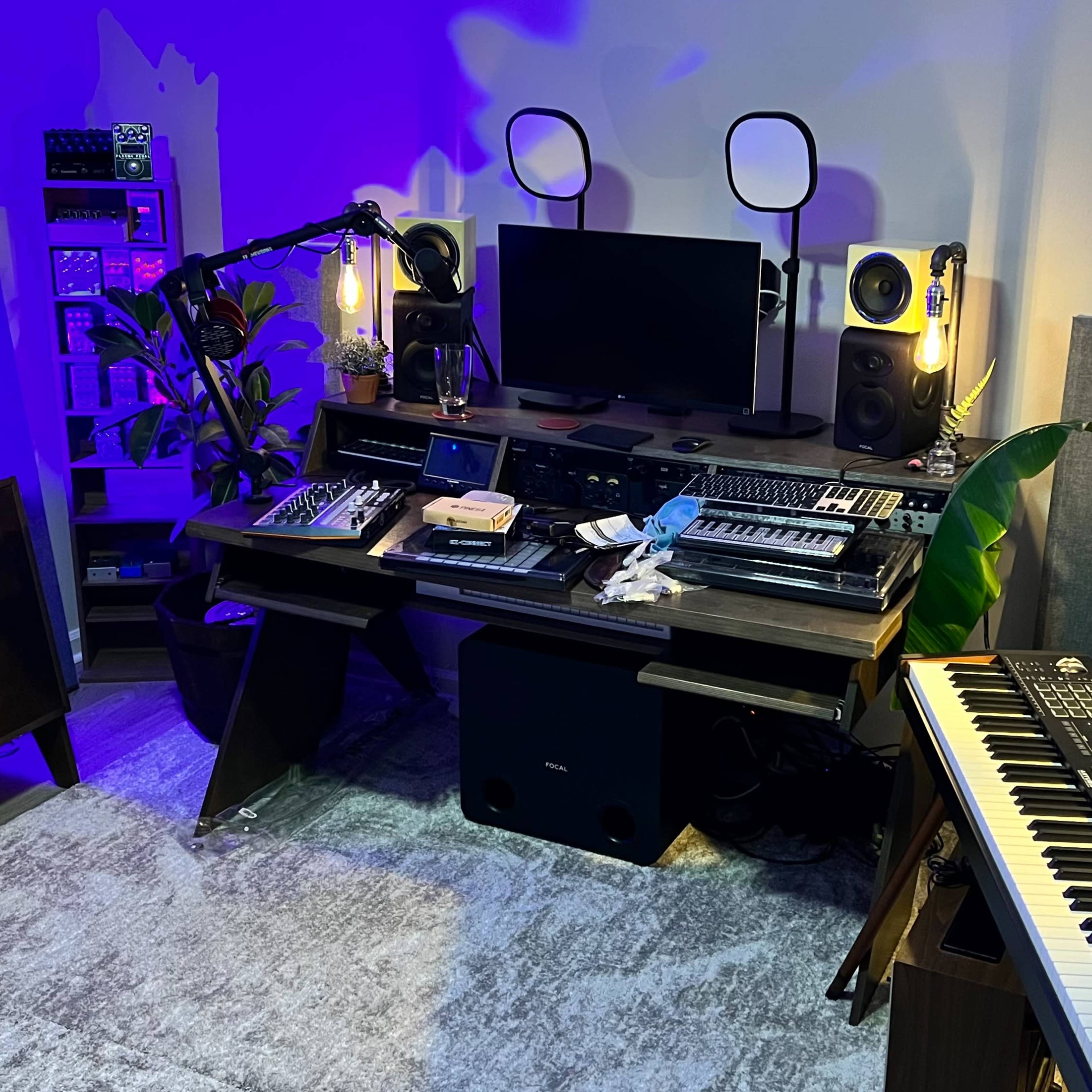 A music production studio with synthesizers, a computer monitor, and various audio equipment arranged around a central desk, with plants and ambient lighting in the background.