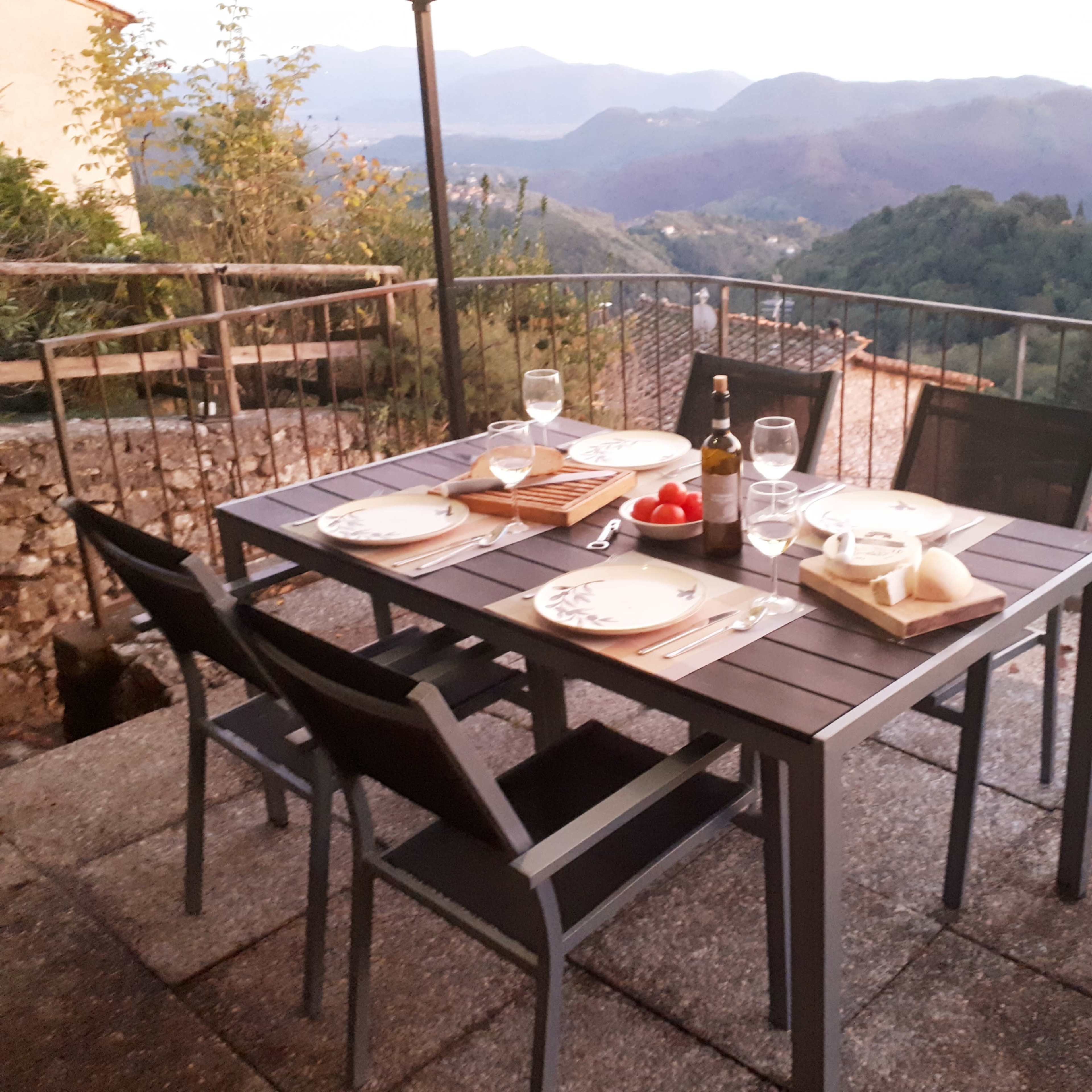 A dining table set with plates, utensils, glasses, and food overlooks a mountainous landscape during twilight.