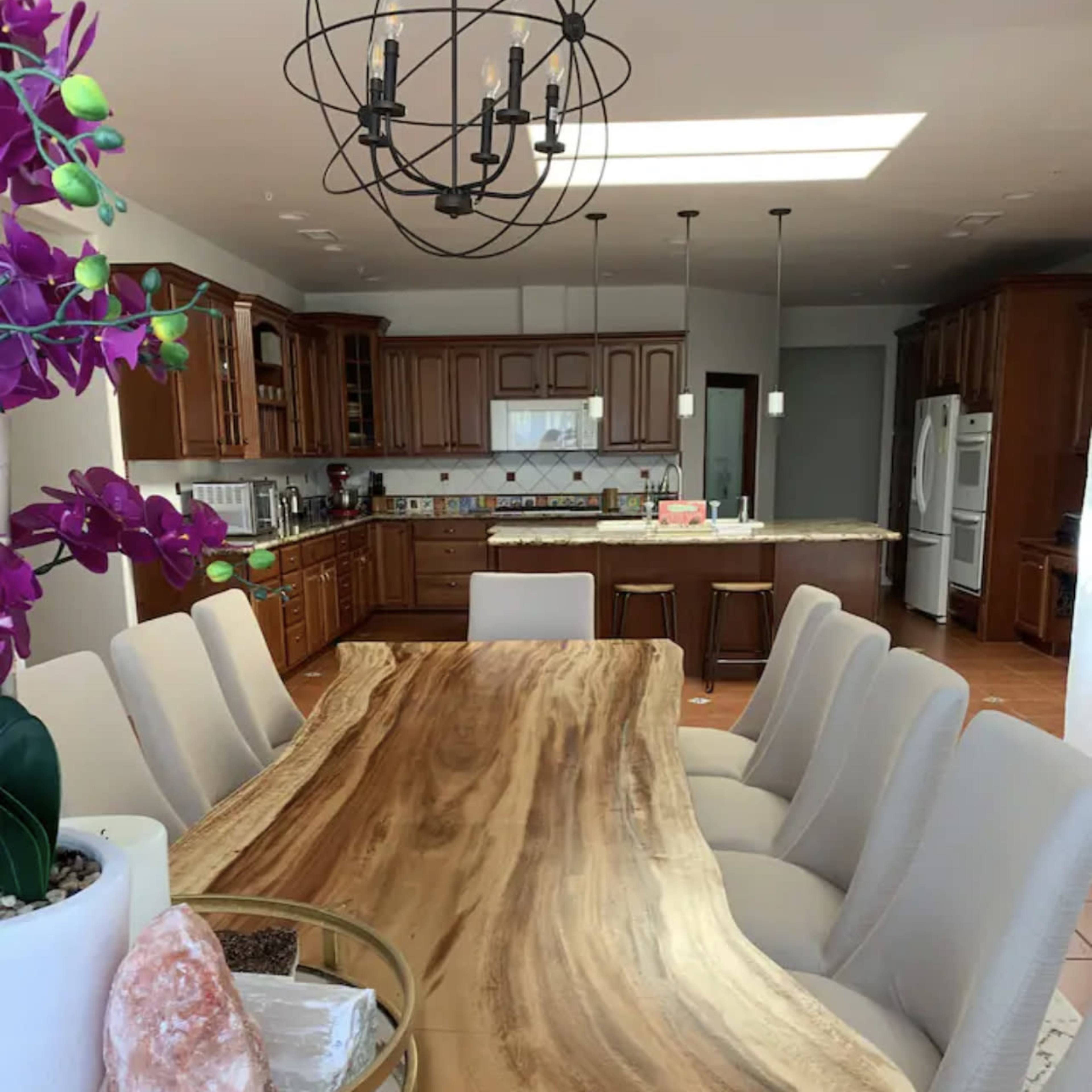 A large, natural wood dining table with modern white chairs is set in a spacious kitchen area featuring dark cabinetry and an open layout.