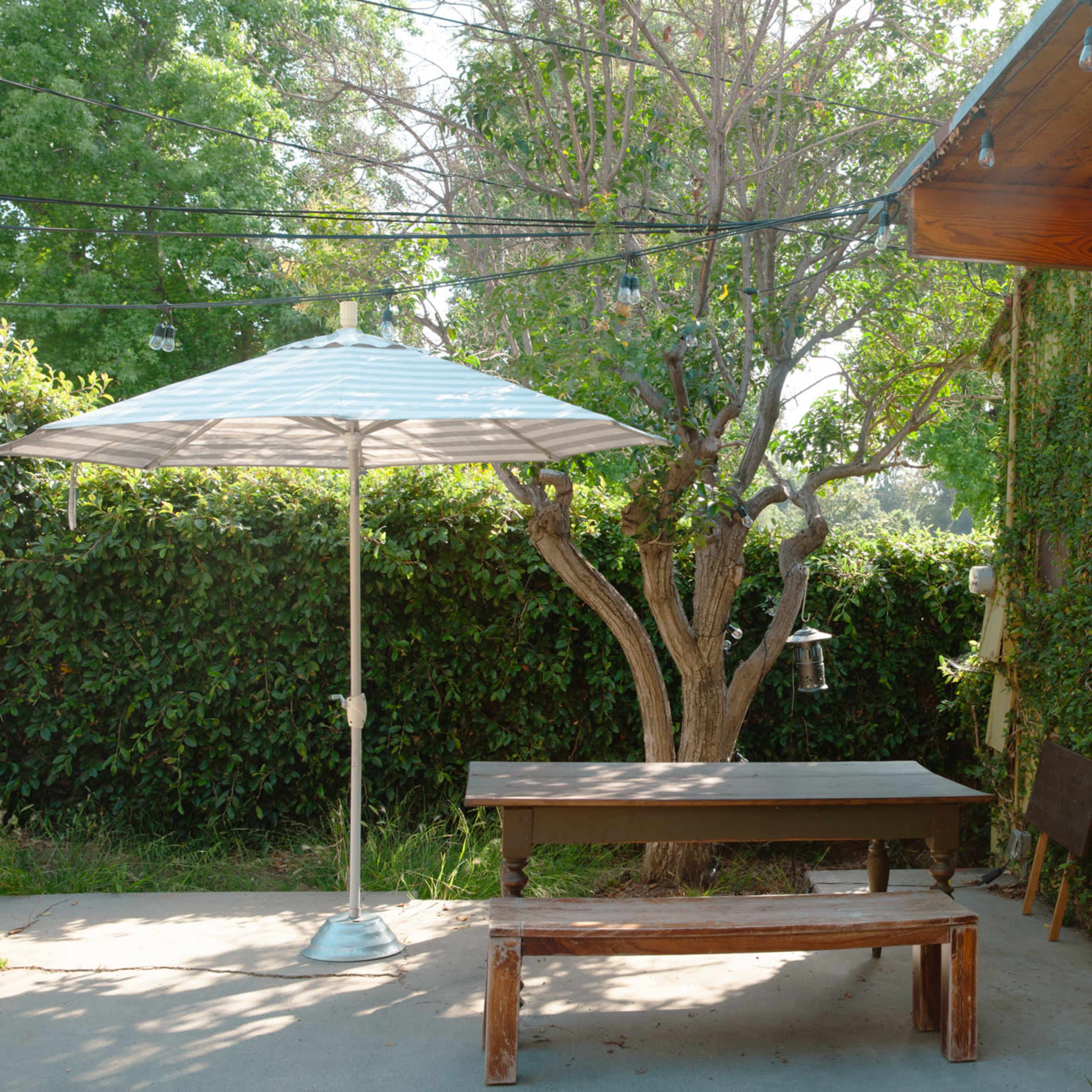A wooden table and bench are positioned beneath a large umbrella in a shaded outdoor area surrounded by green foliage and a glass wall.