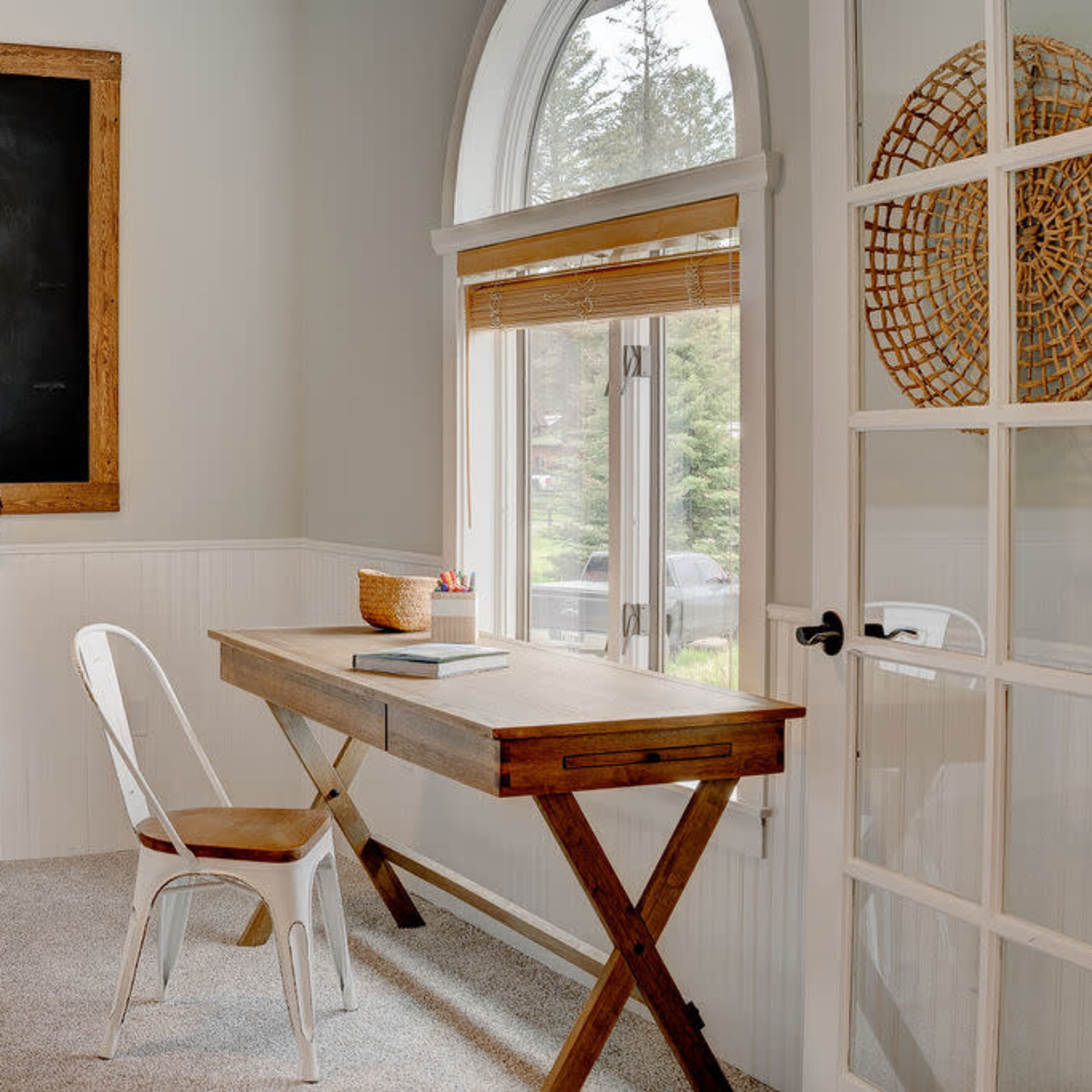 A minimalist home office with a wooden desk, a white chair, a chalkboard on the wall, and a large window with natural light.