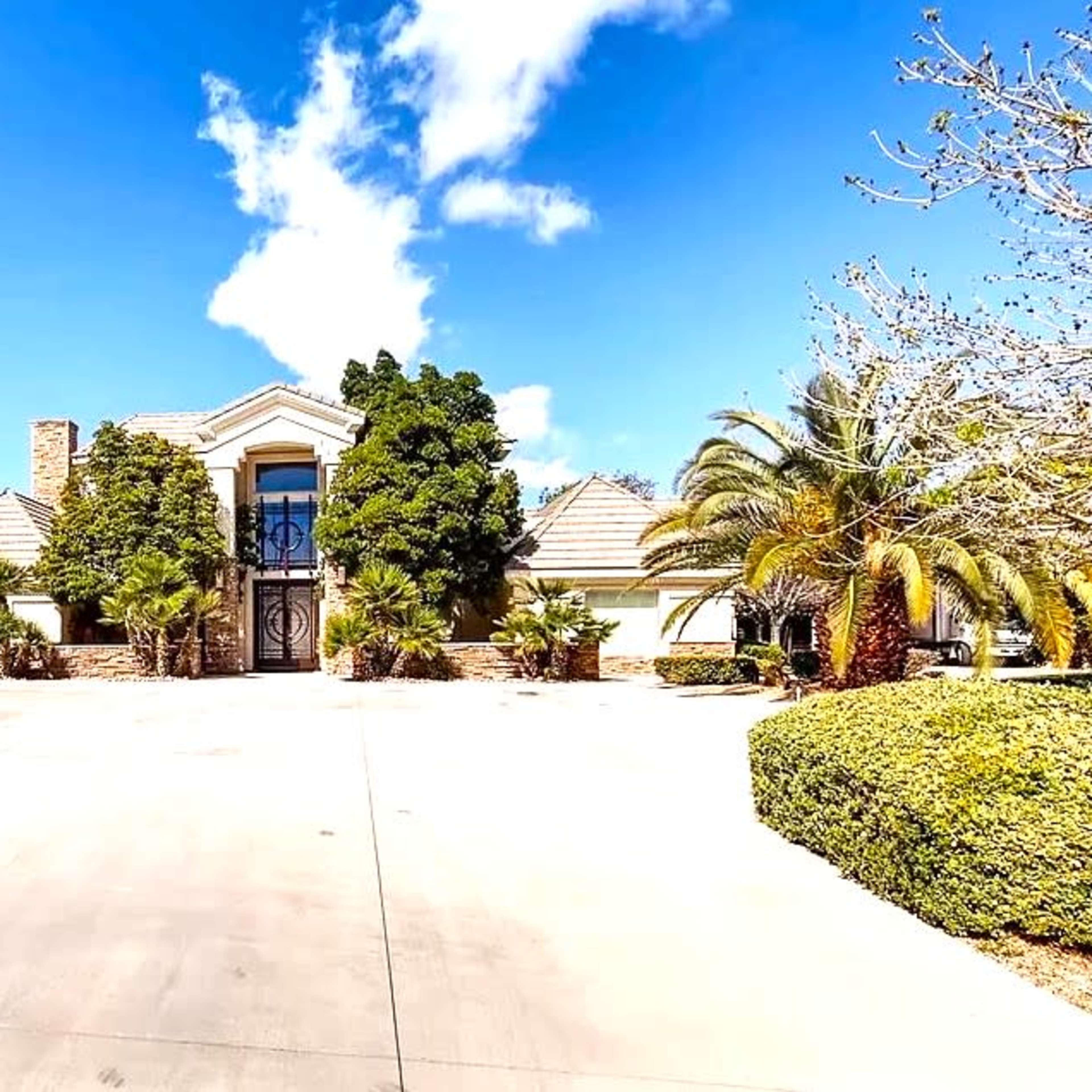 A large, modern house with a stone entrance is surrounded by well-manicured landscaping and palm trees, set against a clear blue sky.