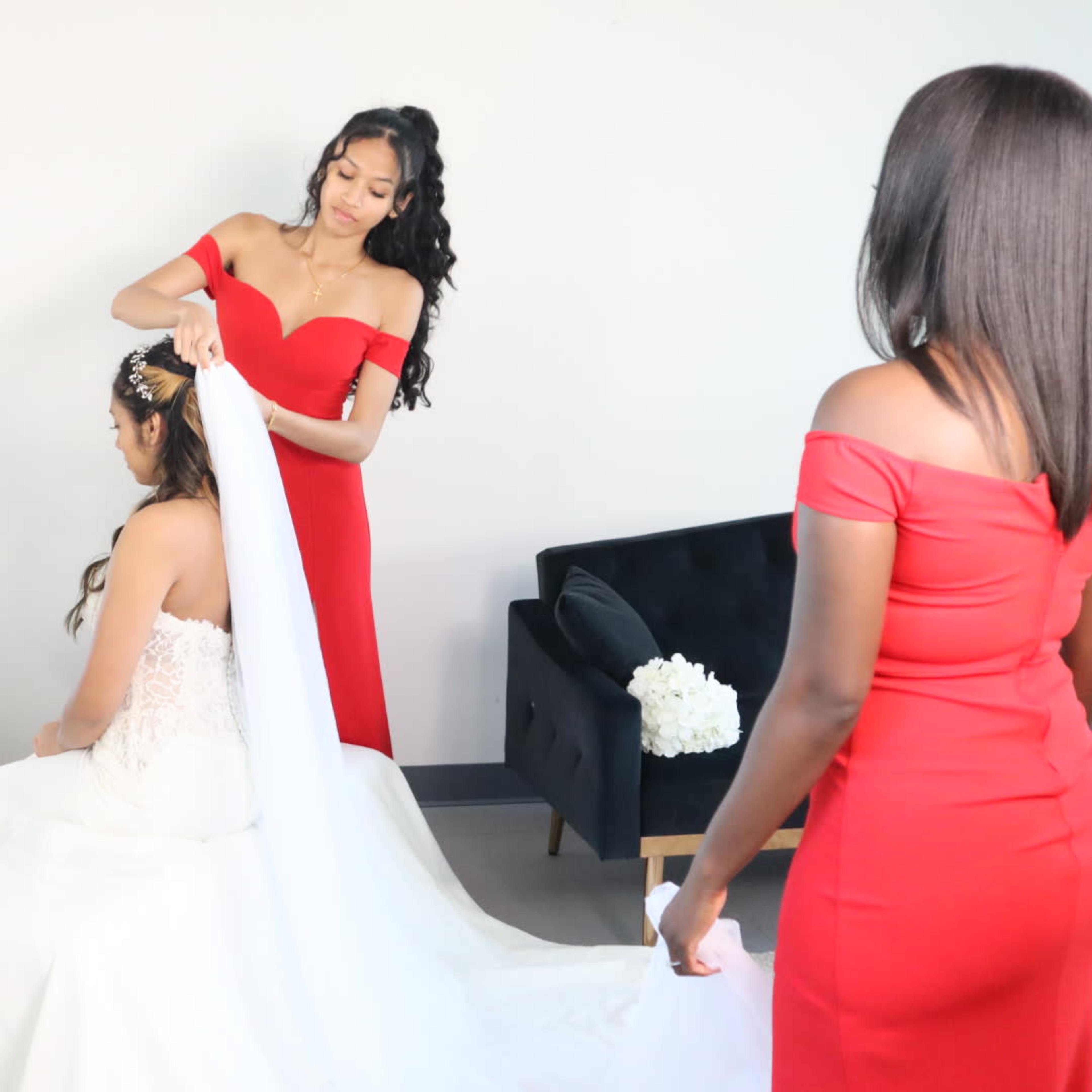 A woman in a white wedding dress sits in front of a mirror while another woman adjusts her veil and a third woman stands nearby, both wearing red dresses.
