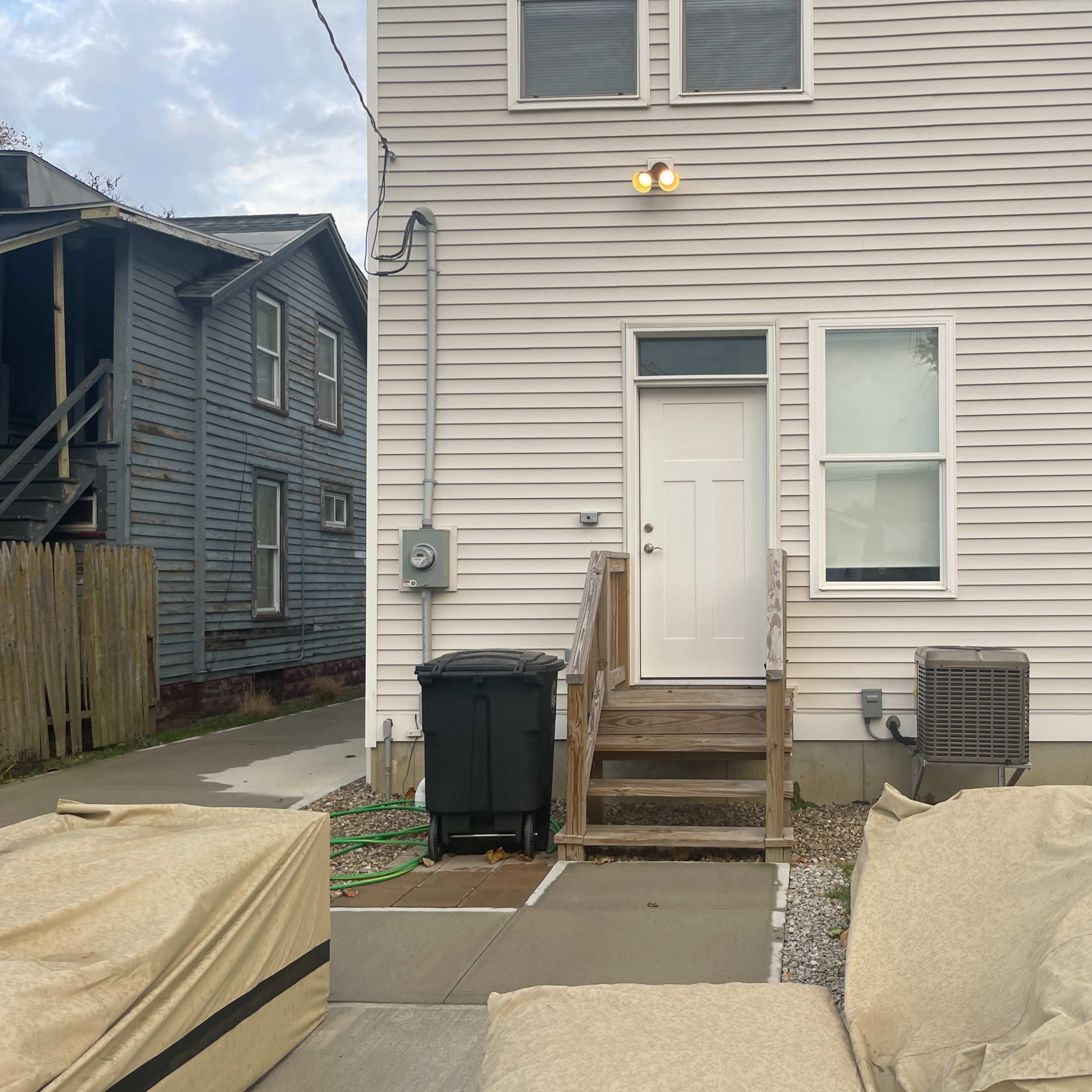 The back entrance of a two-story house, with stairs leading up to a white door, surrounded by a paved area and outdoor furniture covered with tan tarps.