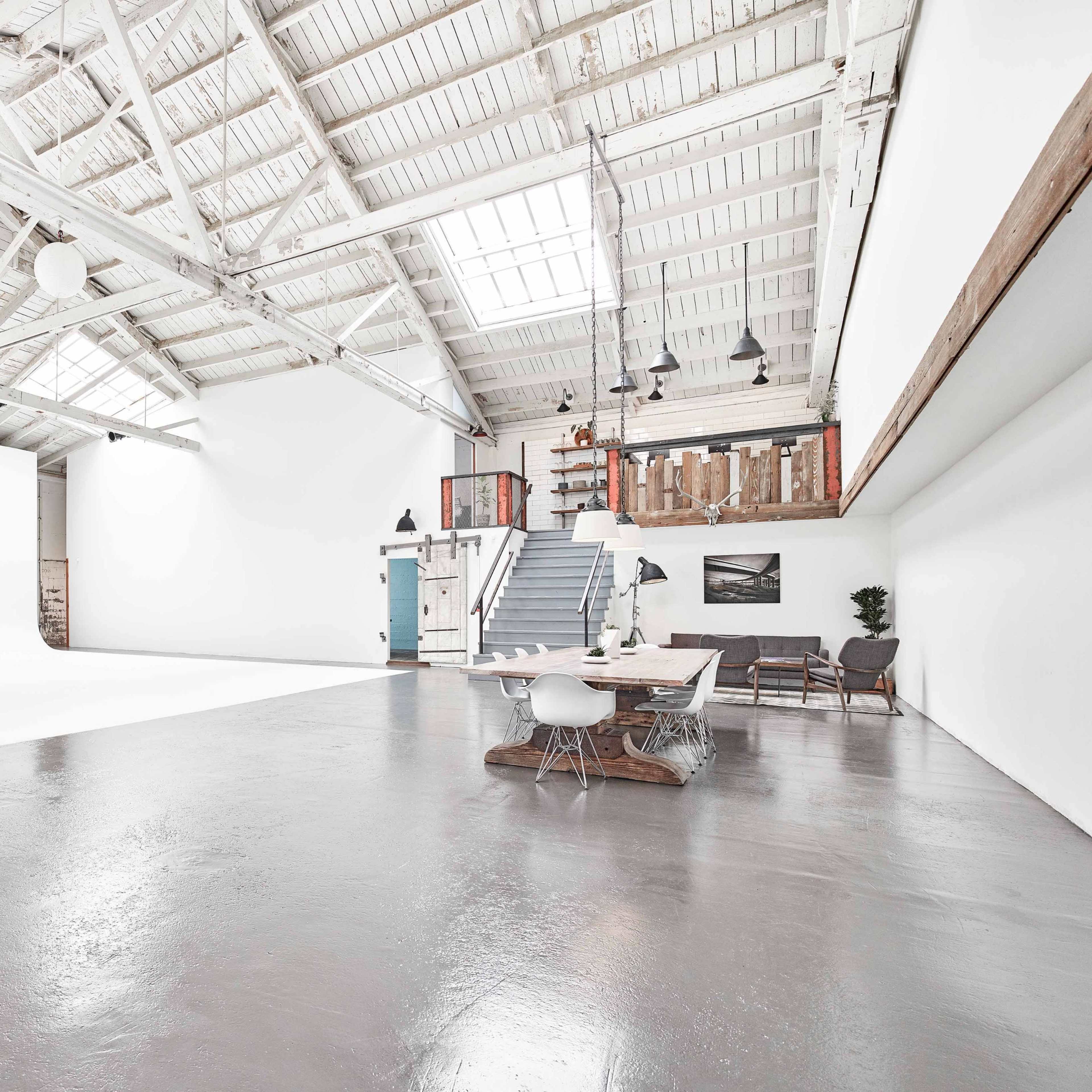A spacious, industrial-style interior with high ceilings, white walls, and a staircase leading to an upper level.