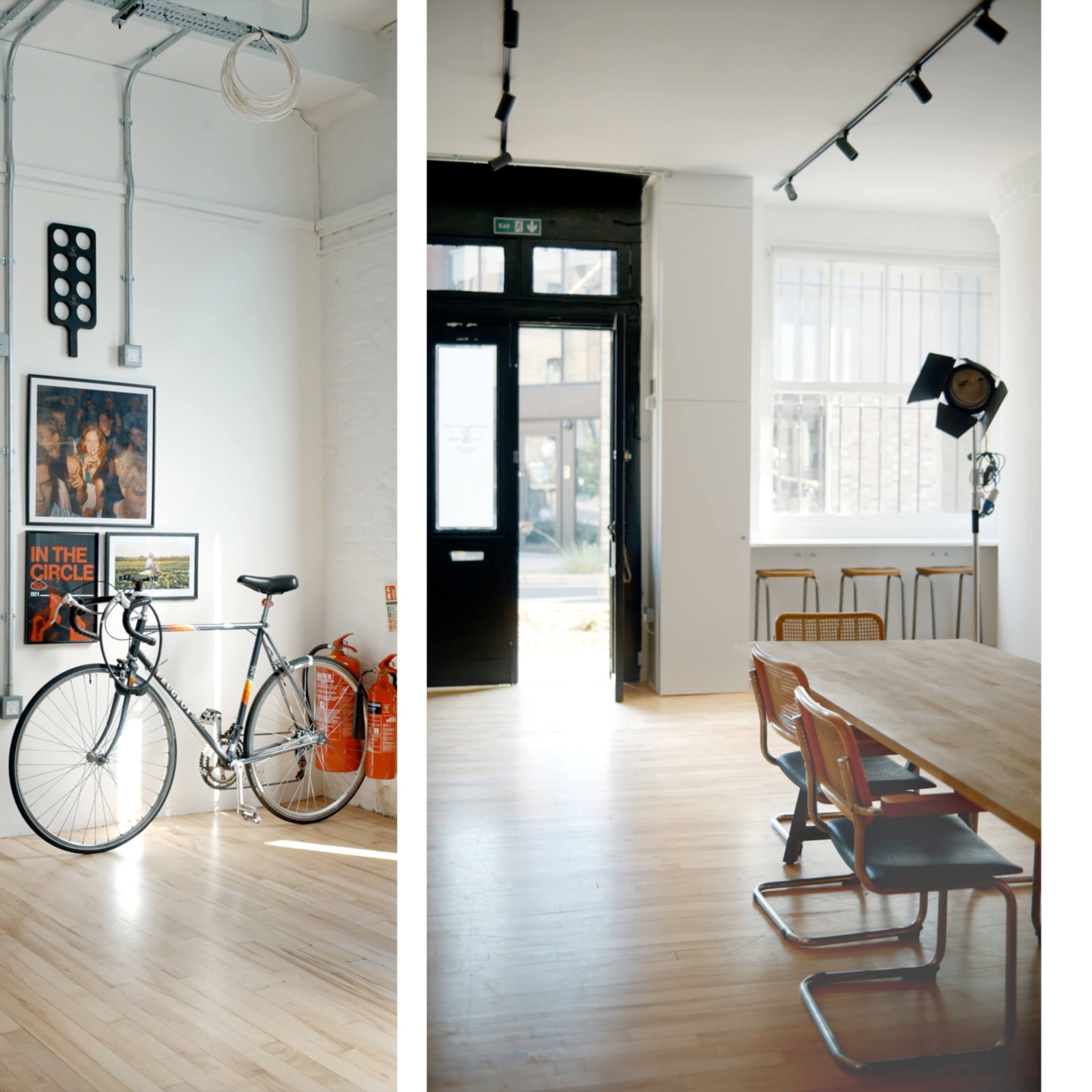A modern interior space featuring a bicycle against a wall with framed art on one side, and a dining area with a large wooden table and metal chairs on the other.