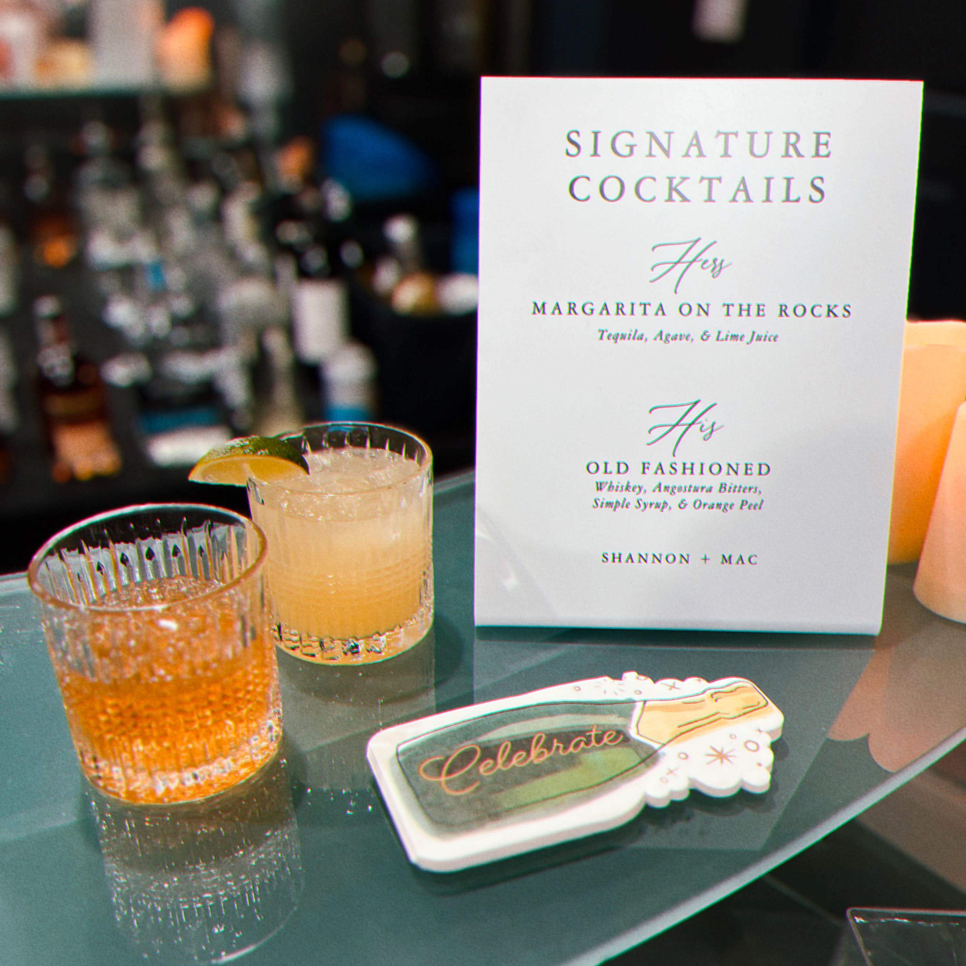 Two cocktails on a glass surface alongside a menu featuring signature cocktails for a wedding celebration.