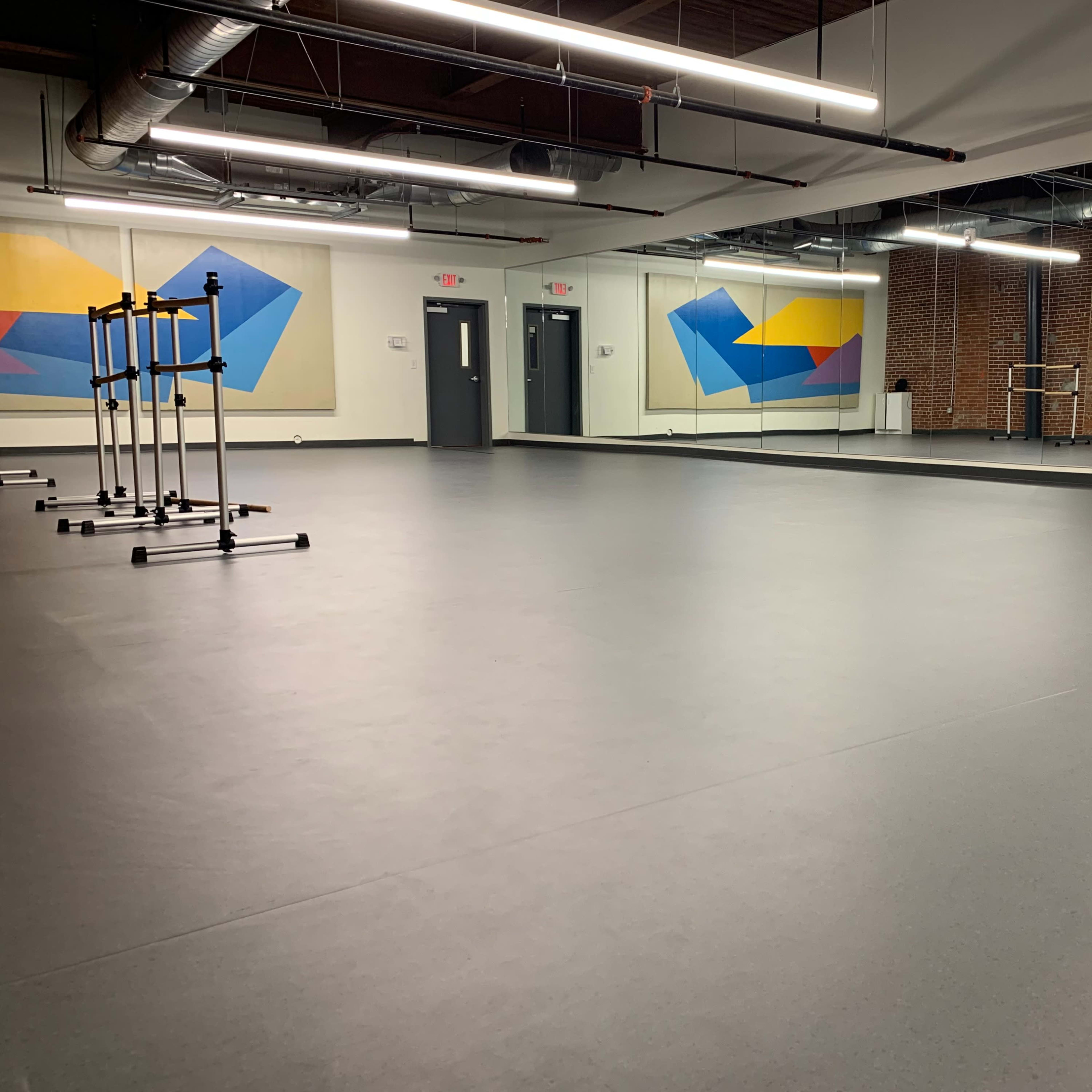 A spacious dance studio with a smooth black floor, mirrored walls, and colorful abstract artwork on the back wall.