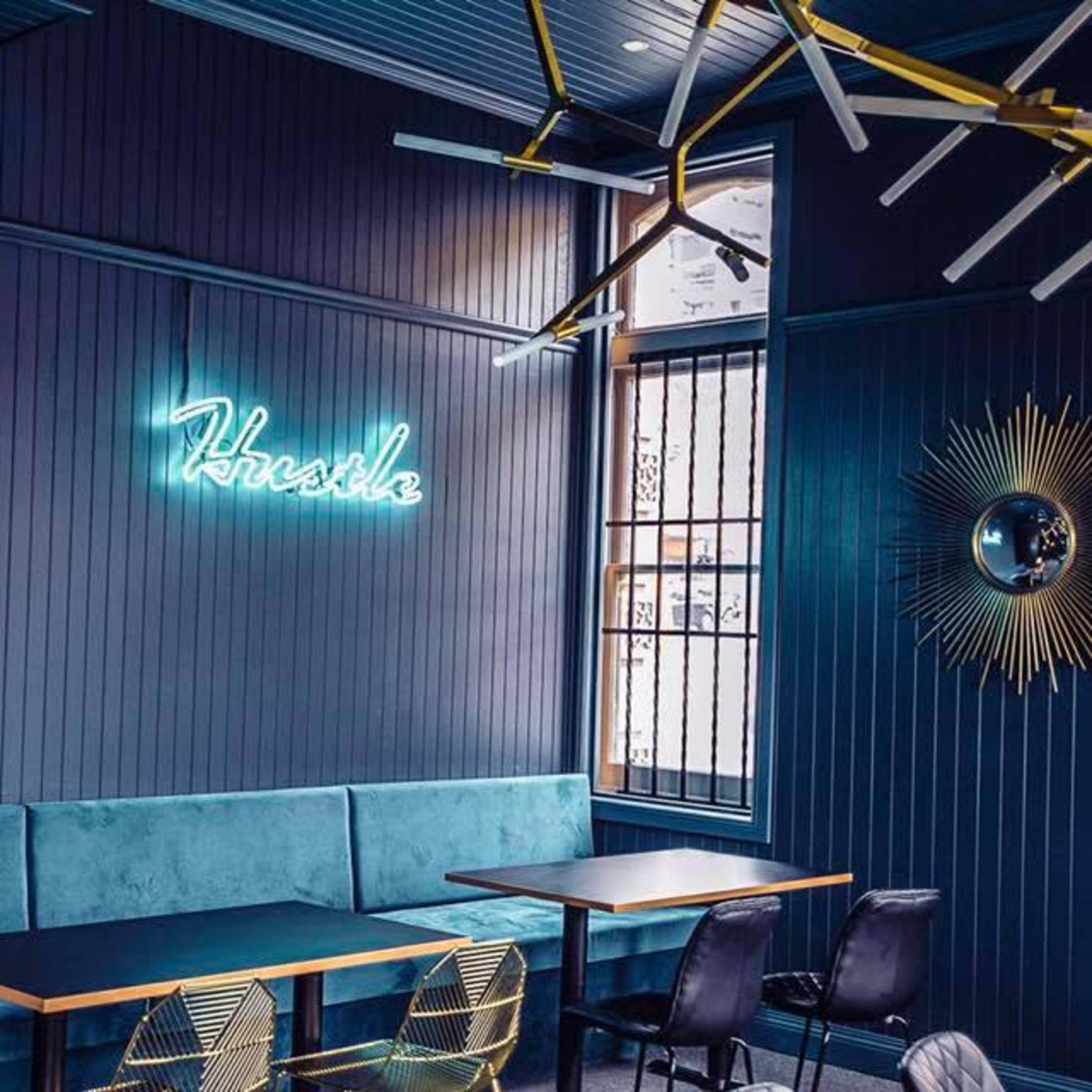 A stylish café interior featuring a blue accent wall with a neon sign that reads "Hustle," a plush seating area, and modern decor elements.