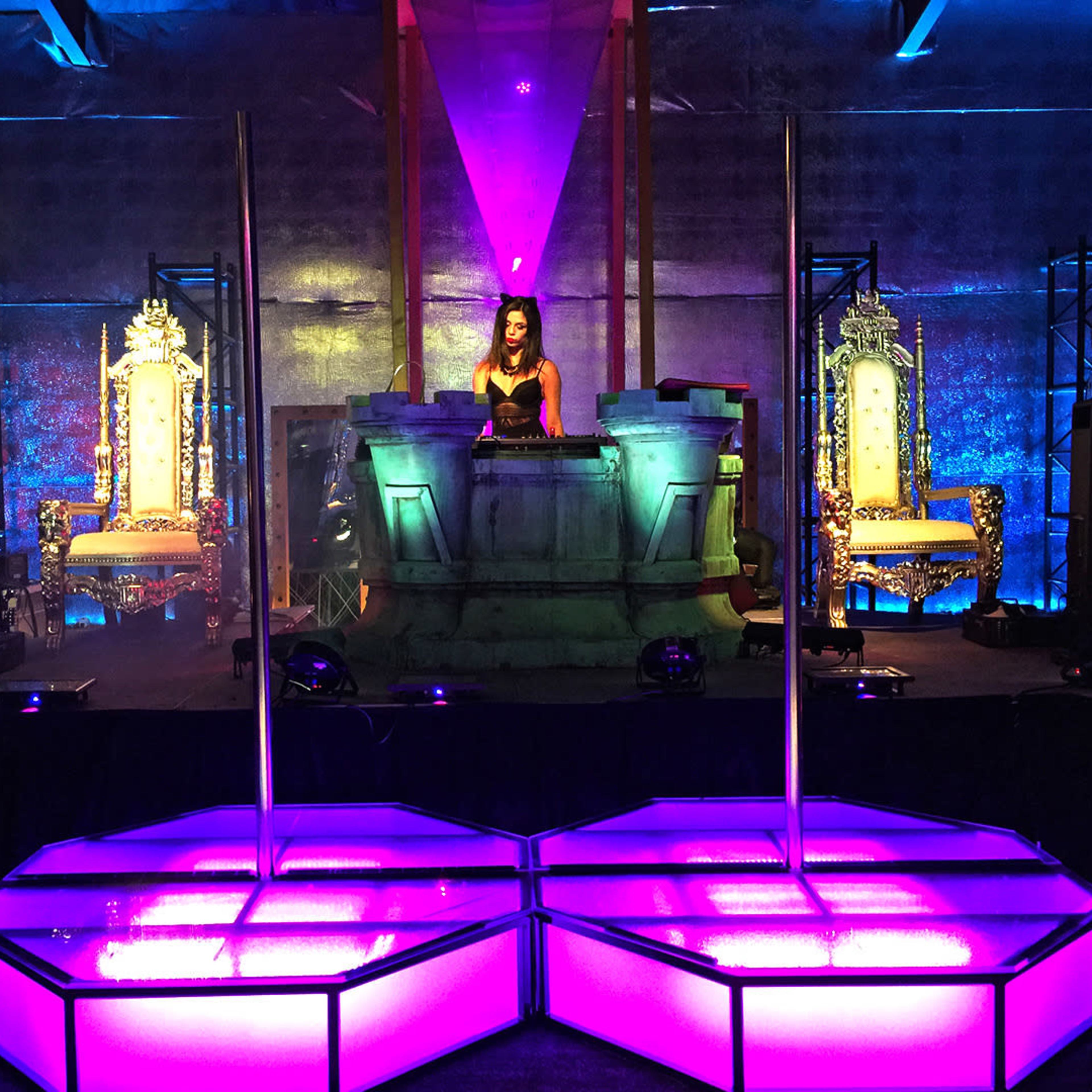 A DJ performs on stage in front of ornate thrones, surrounded by colorful lighting and large speakers.