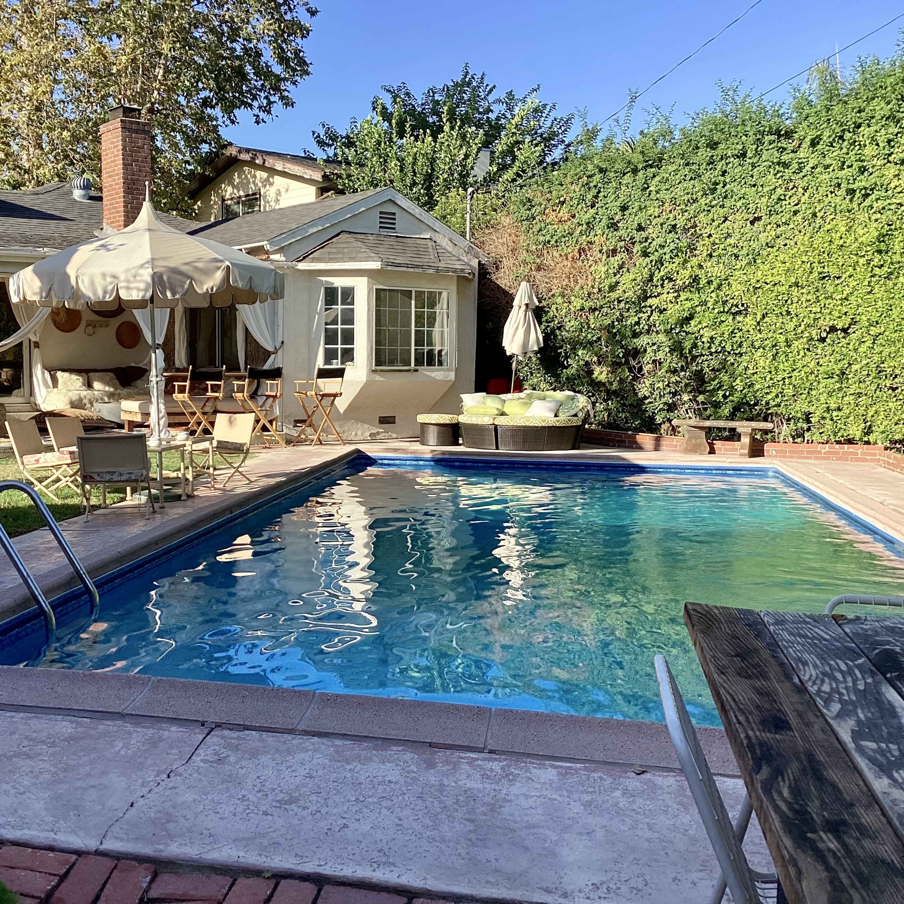 A residential backyard with a swimming pool, a covered seating area, and a house in the background surrounded by green foliage.