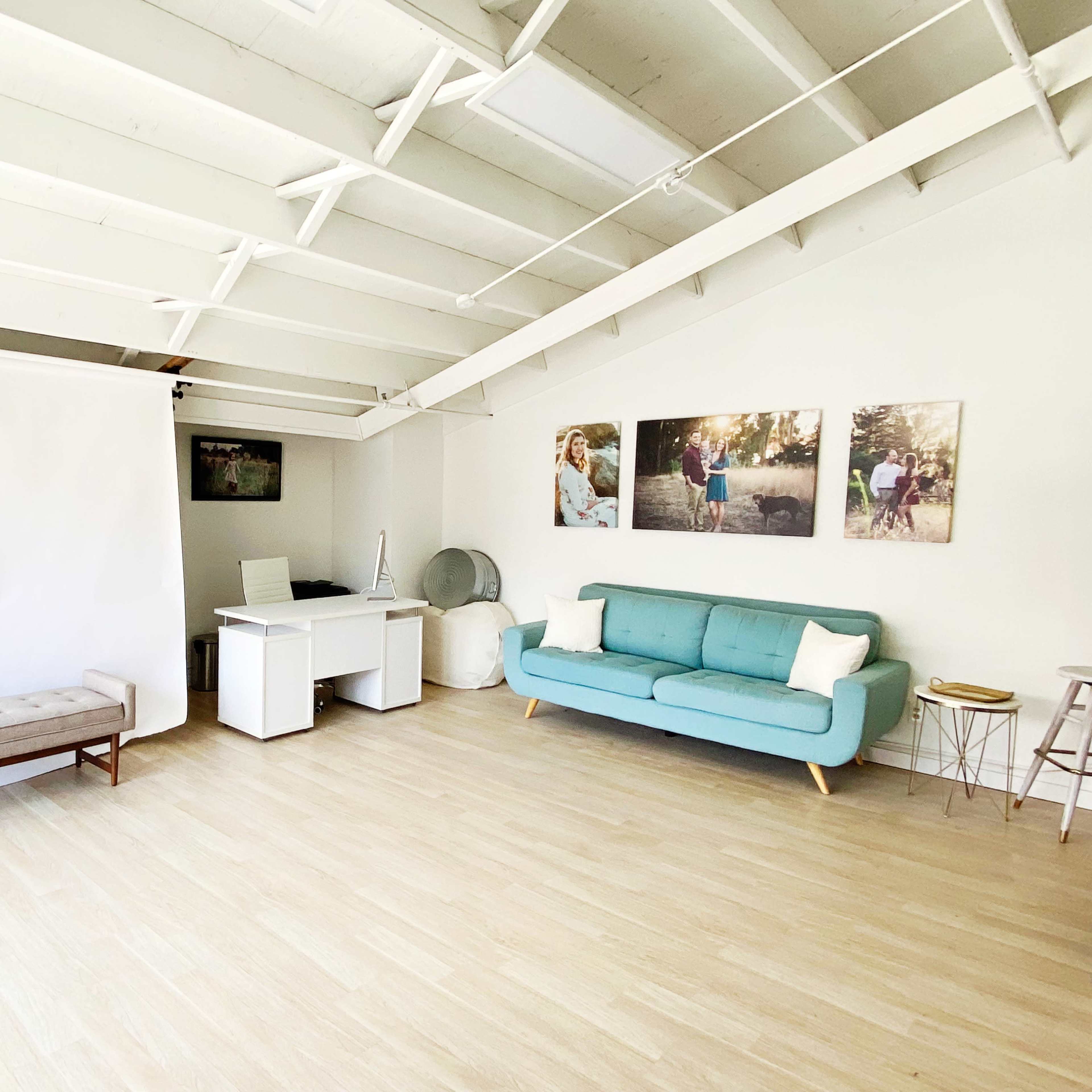 A bright, spacious room with a light wood floor, a teal sofa, a white desk, and two large photographs on the wall.