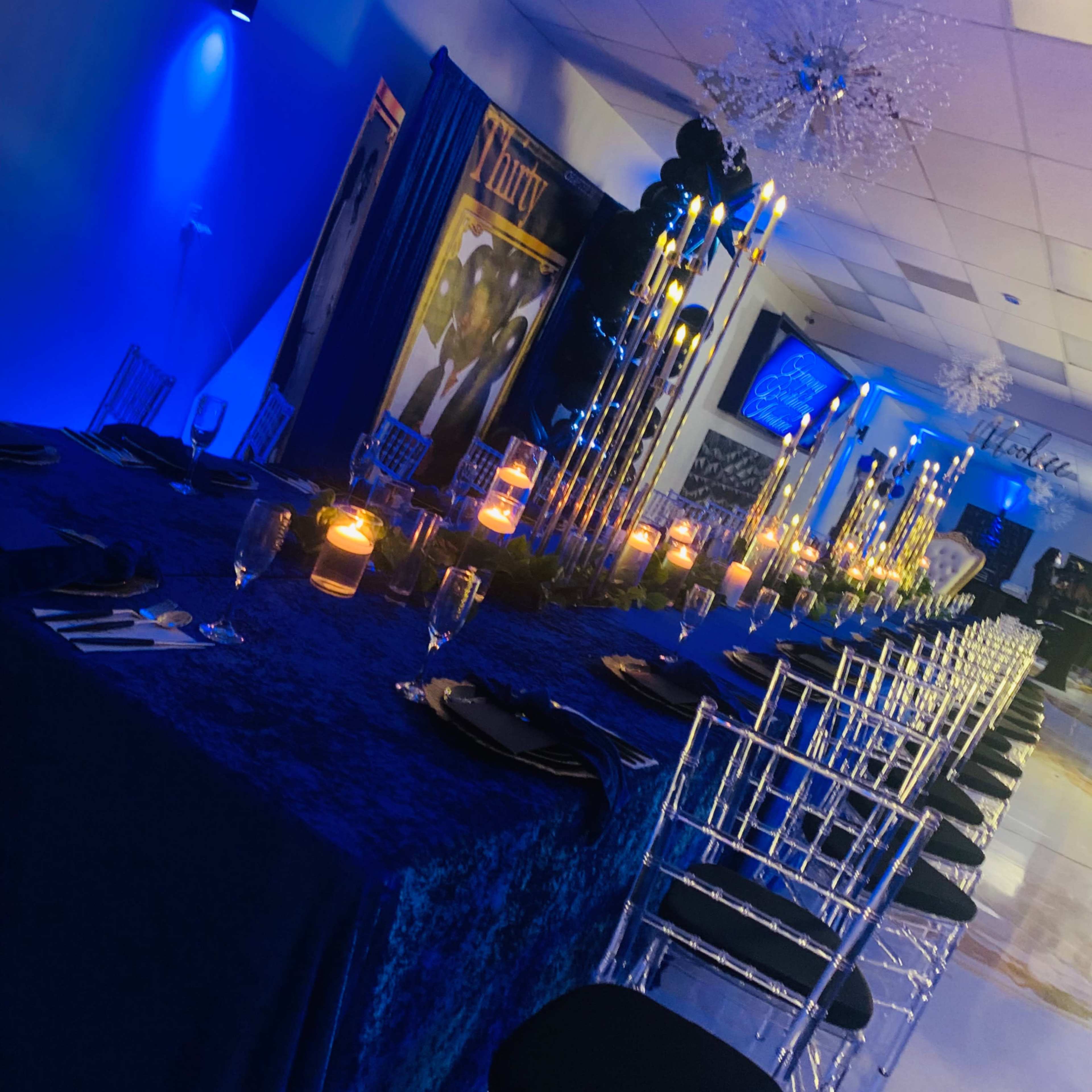 A long dining table is set with candles and elegant black chairs in a dimly lit room with blue lighting and decorative elements.