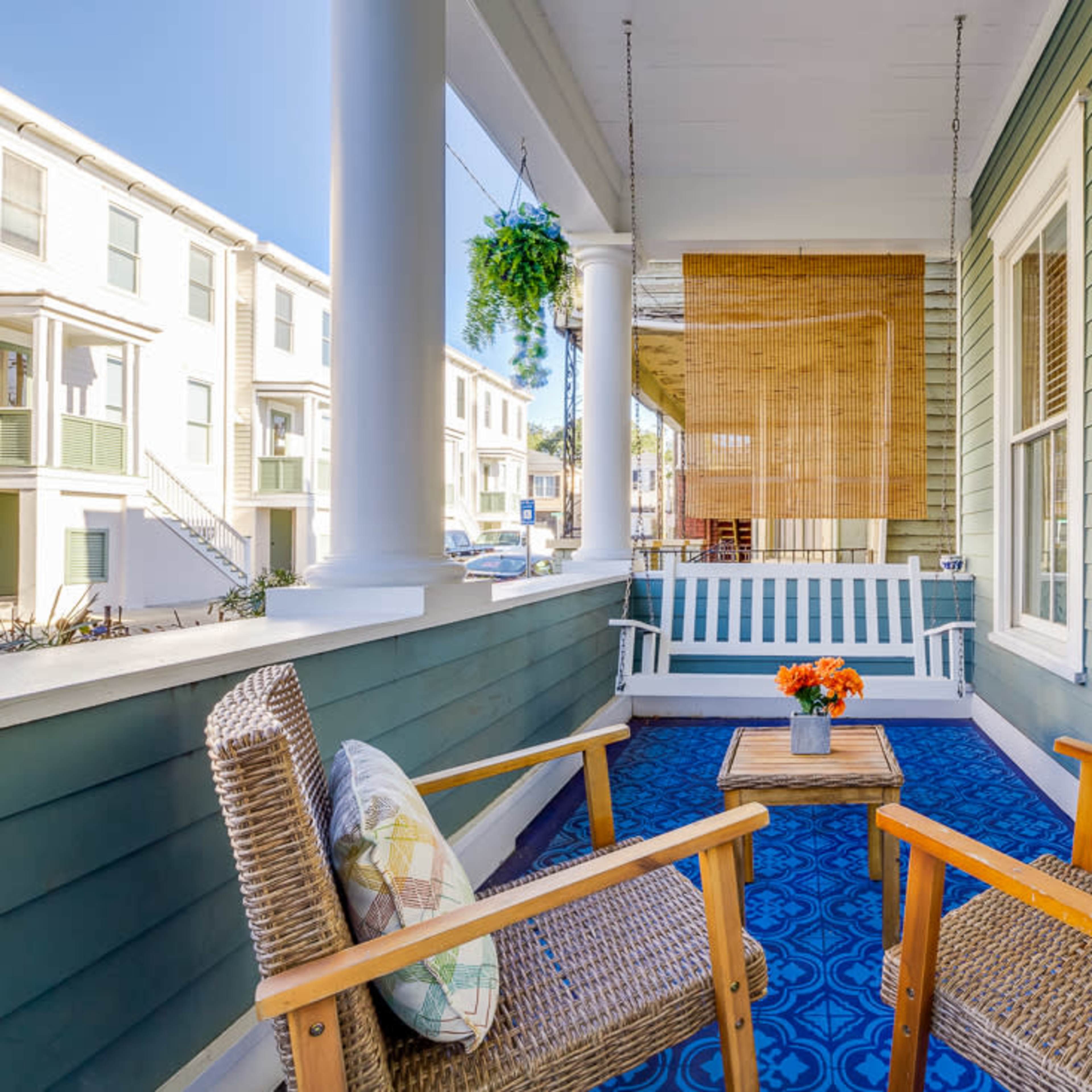 A porch with two wooden chairs, a small table with flowers, and a blue rug, surrounded by green wooden walls and a sunny exterior.