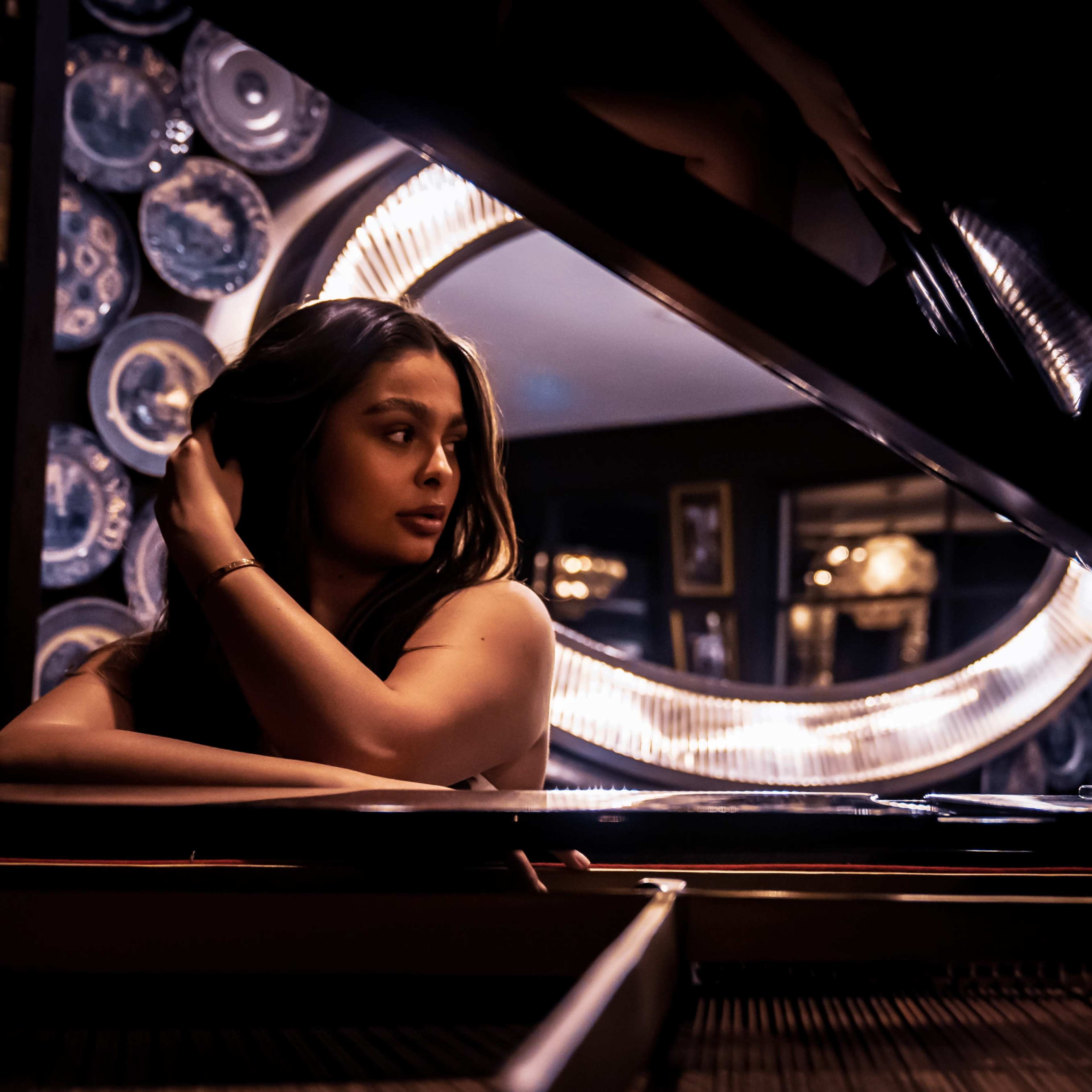 A woman sits at a grand piano in a dimly lit room decorated with circular mirrors and framed pictures.