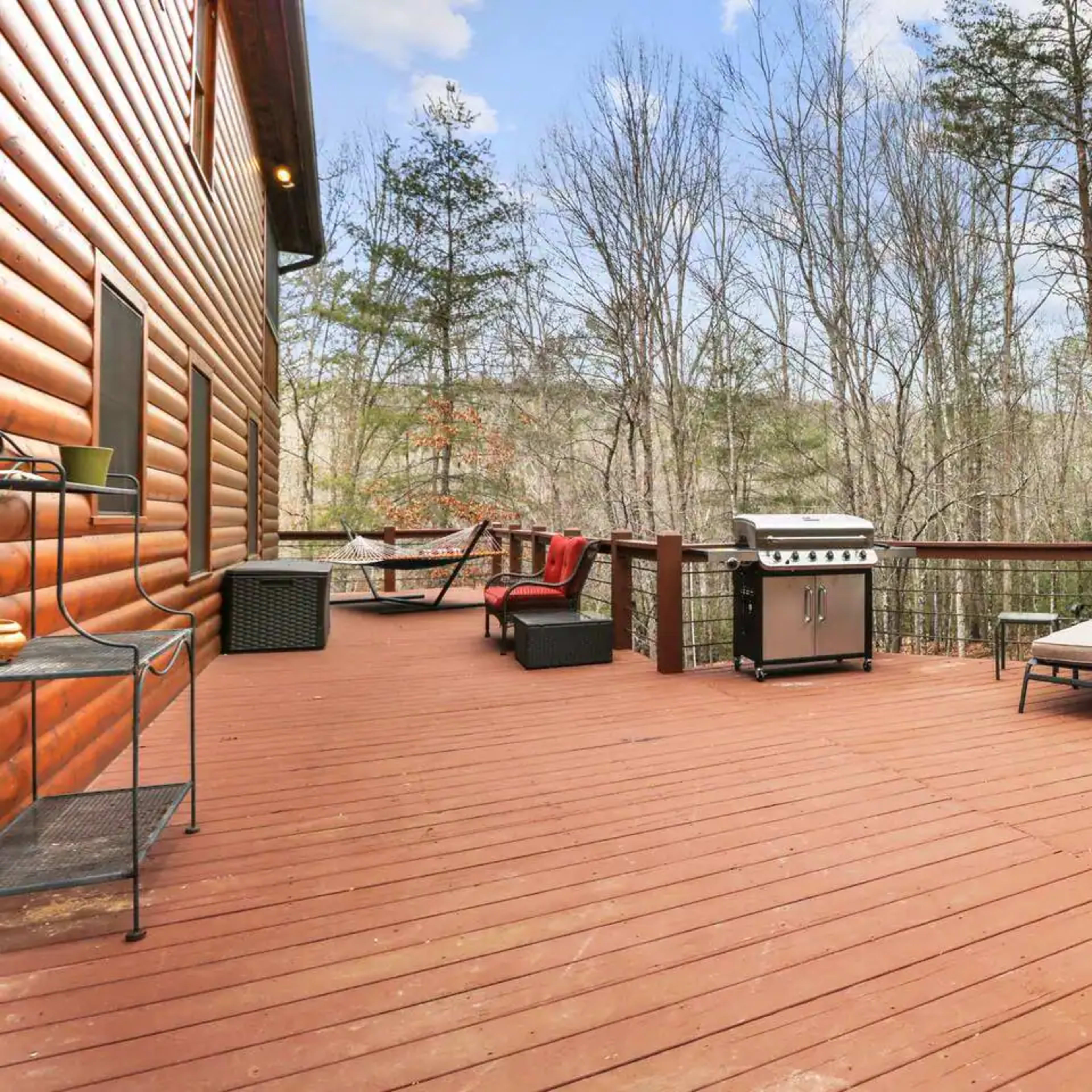 A wooden deck features a grill and two lounge chairs overlooking a wooded area.