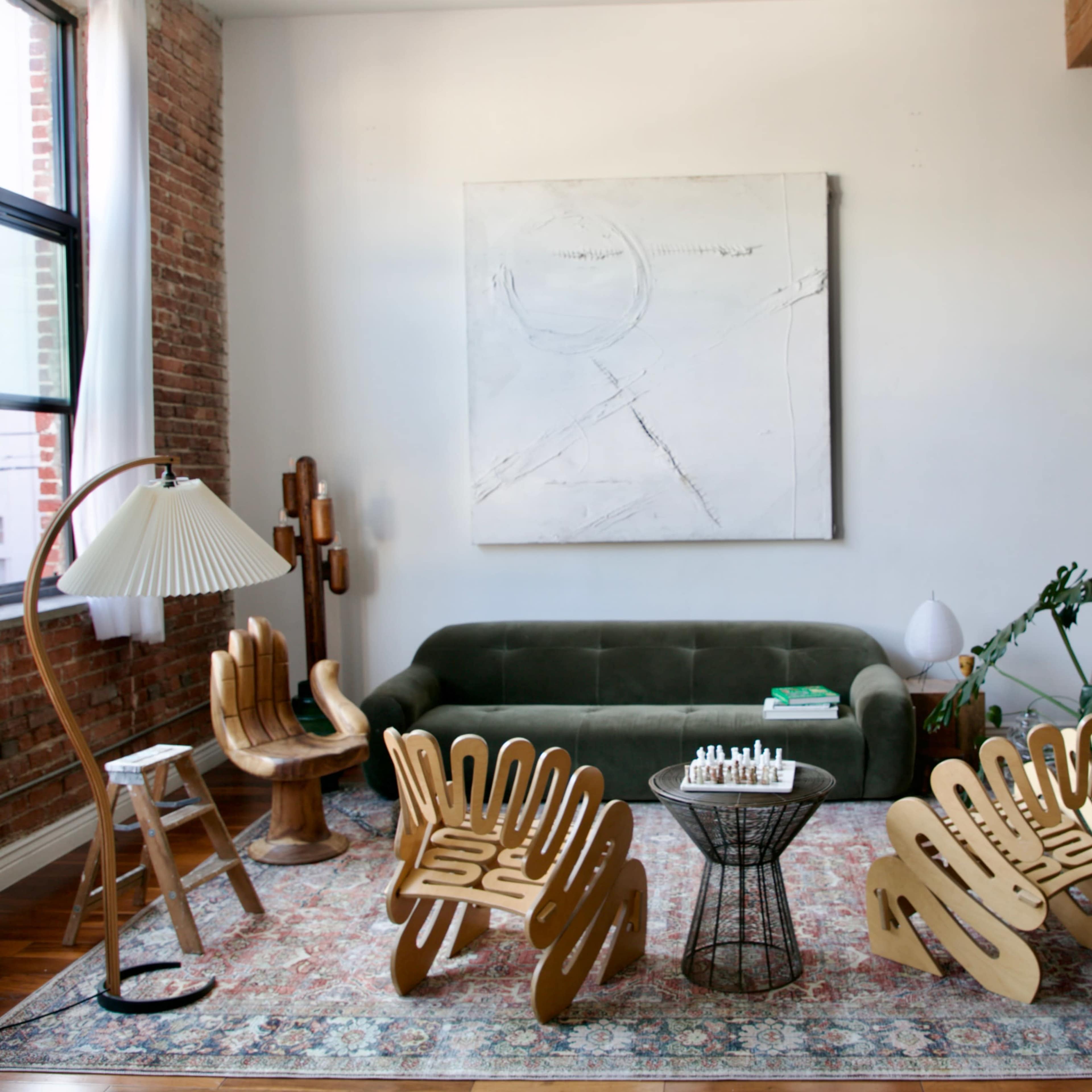 A modern living room featuring a green sofa, unique wooden chairs, a small table with a chess set, and various decorative plants.