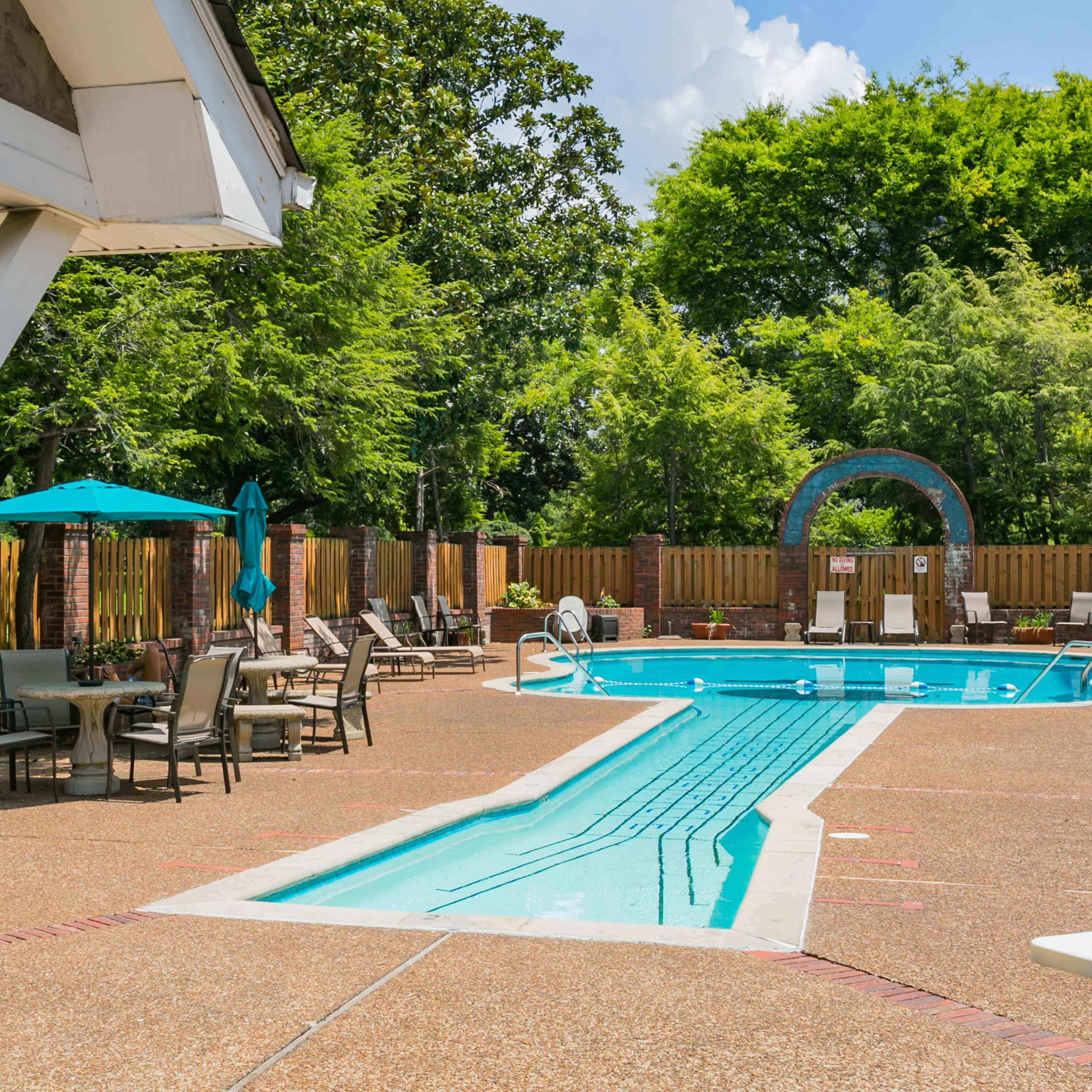 A clear swimming pool surrounded by lounge chairs and patio umbrellas in a landscaped area.