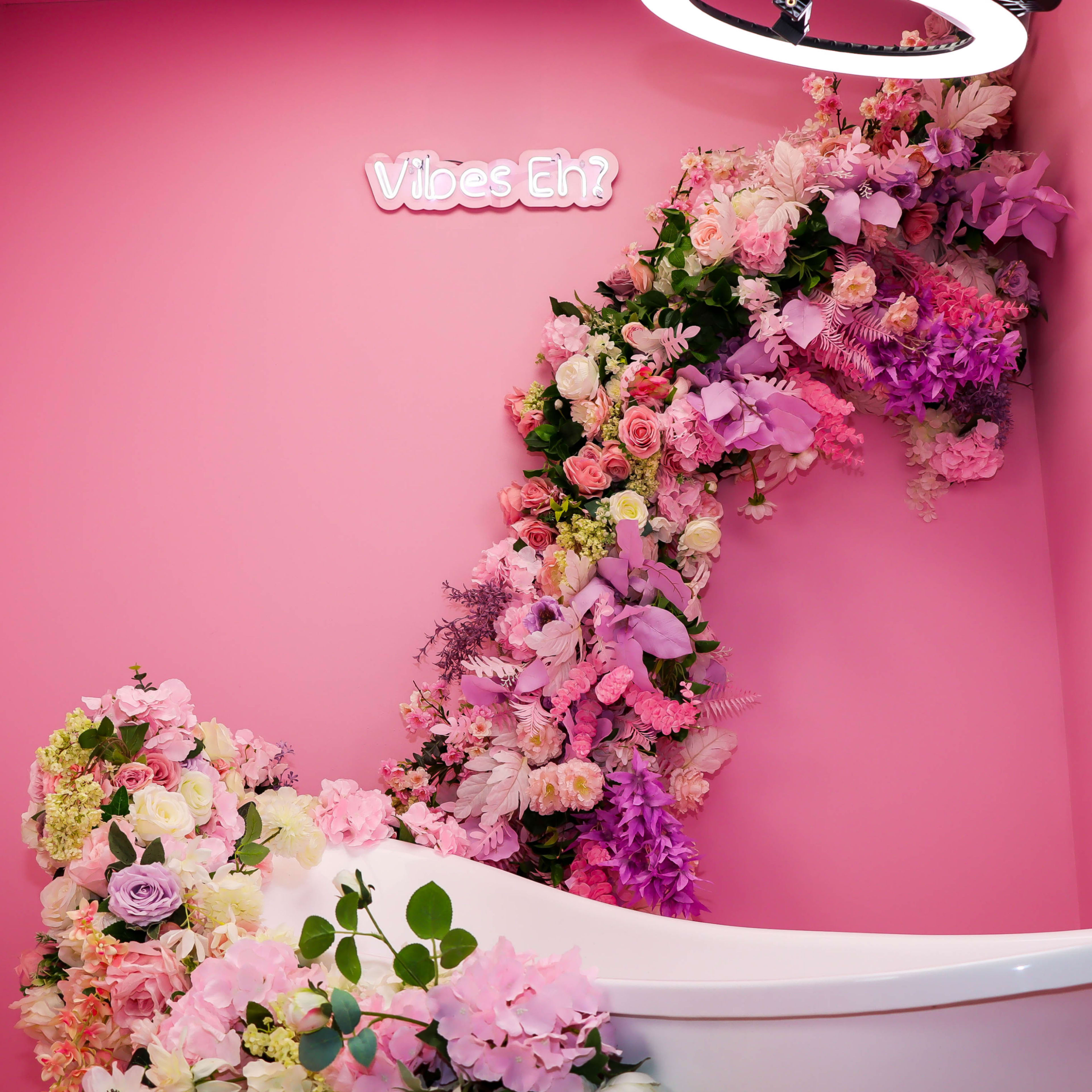 A white bathtub is positioned against a pink wall adorned with a colorful floral arrangement that curves towards a sign reading "Vibes Eh?"
