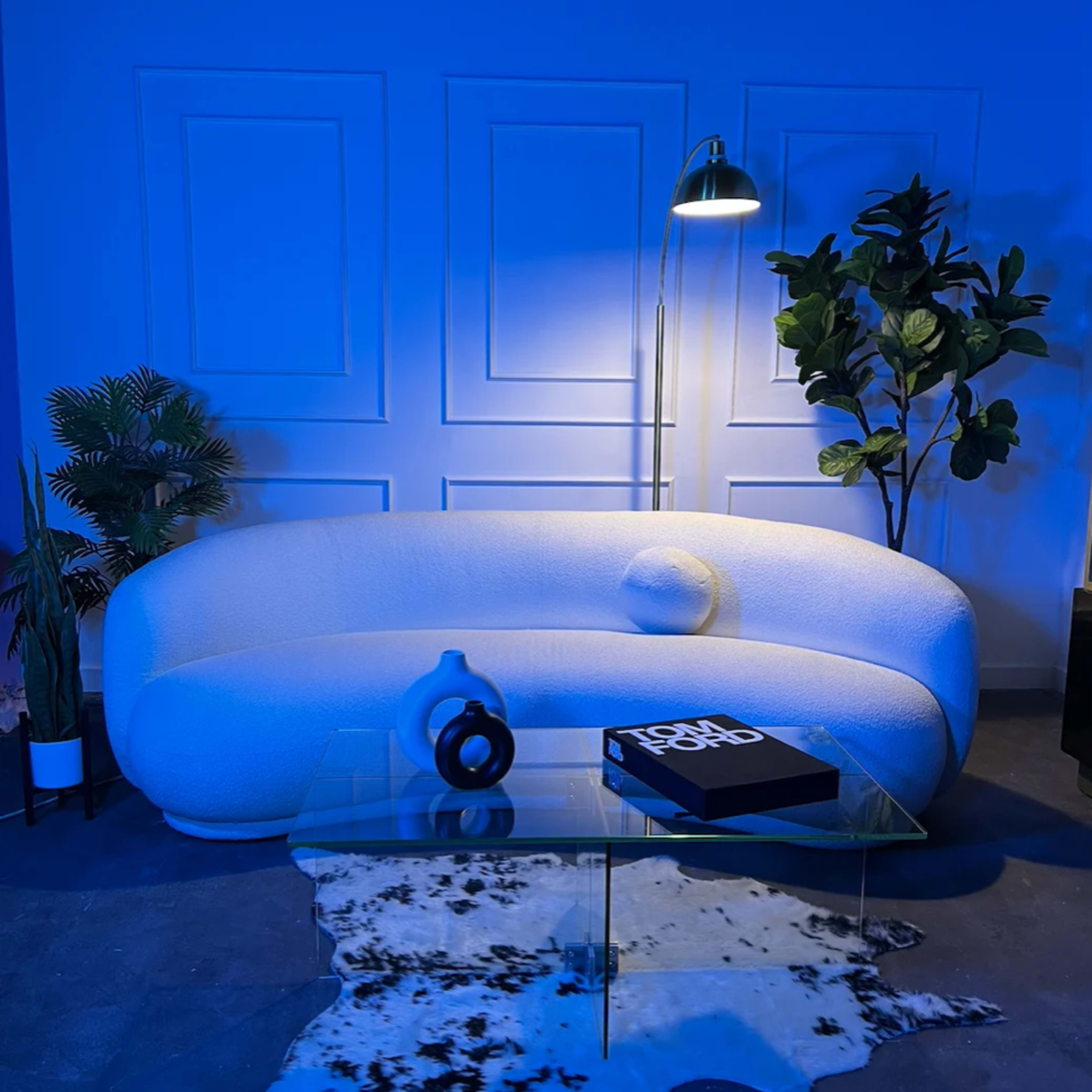 A white, curved sofa is positioned next to a glass coffee table on a cowhide rug, with a floor lamp and a decorative plant in the background, all illuminated by blue lighting.