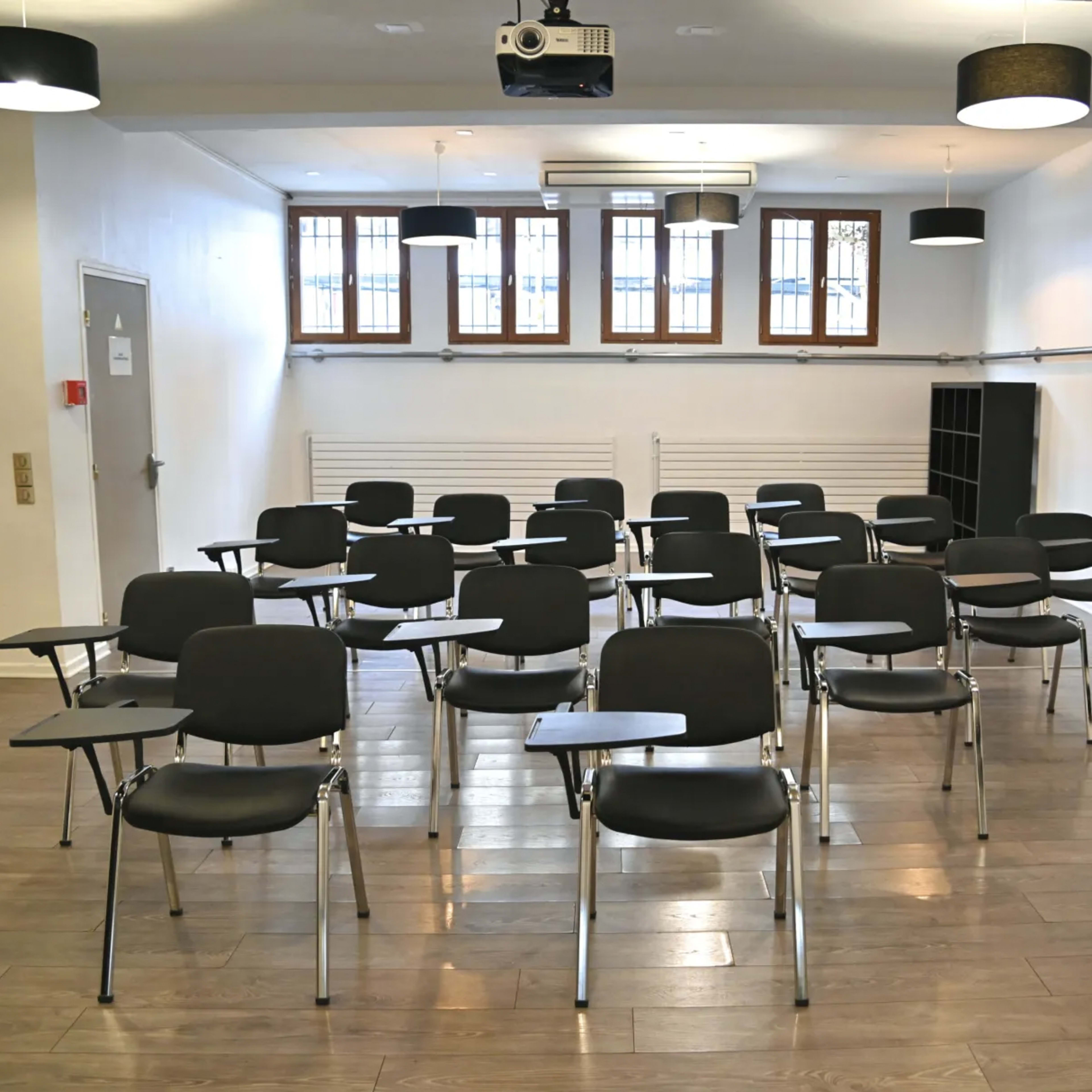 A classroom with rows of black chairs equipped with writing surfaces, arranged neatly on a wooden floor under overhead lighting.