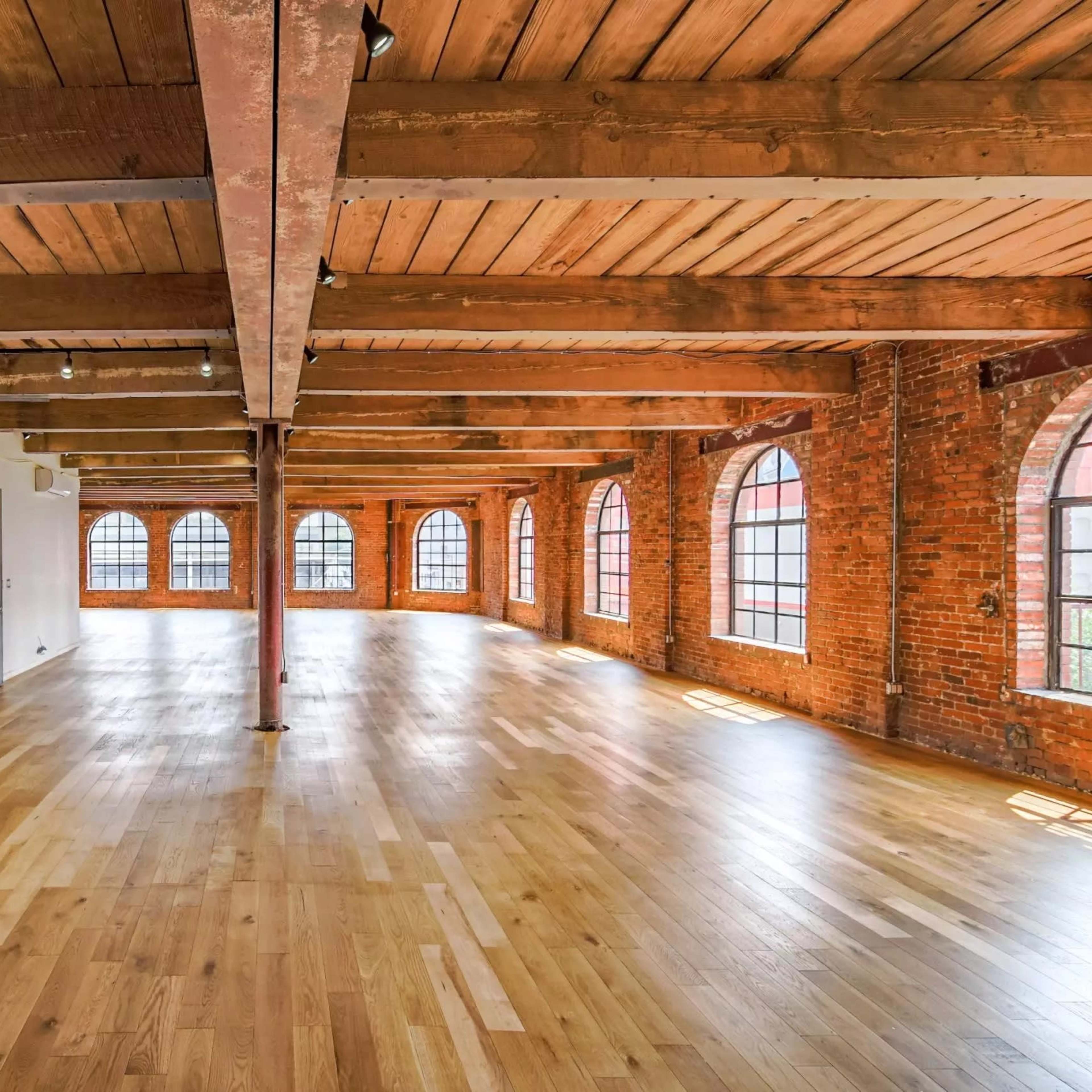 A spacious, open room with wooden floors, exposed brick walls, and large arched windows.