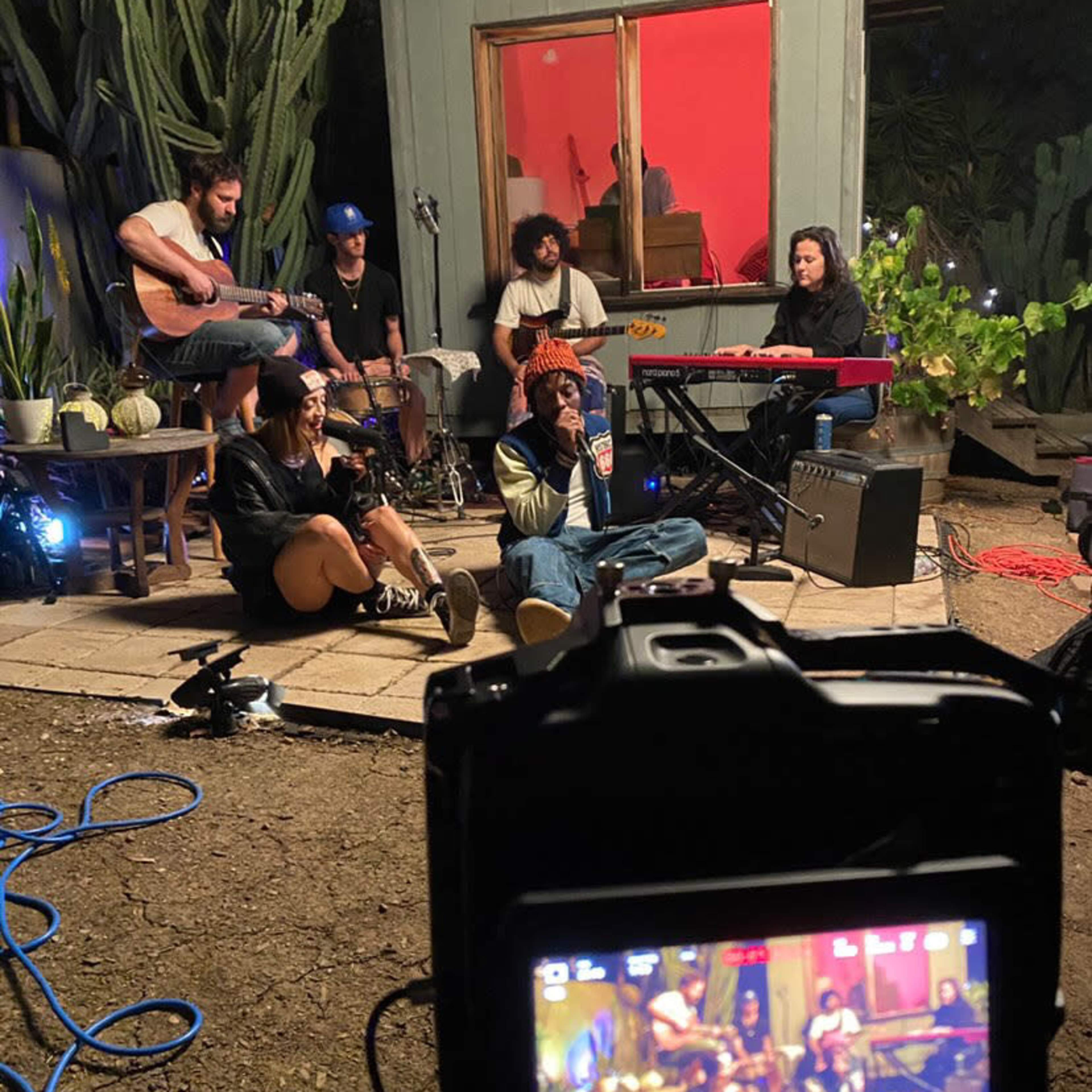 A group of musicians performs together in a backyard setting, with instruments and equipment visible, while some sit on the ground in front of a lit window.