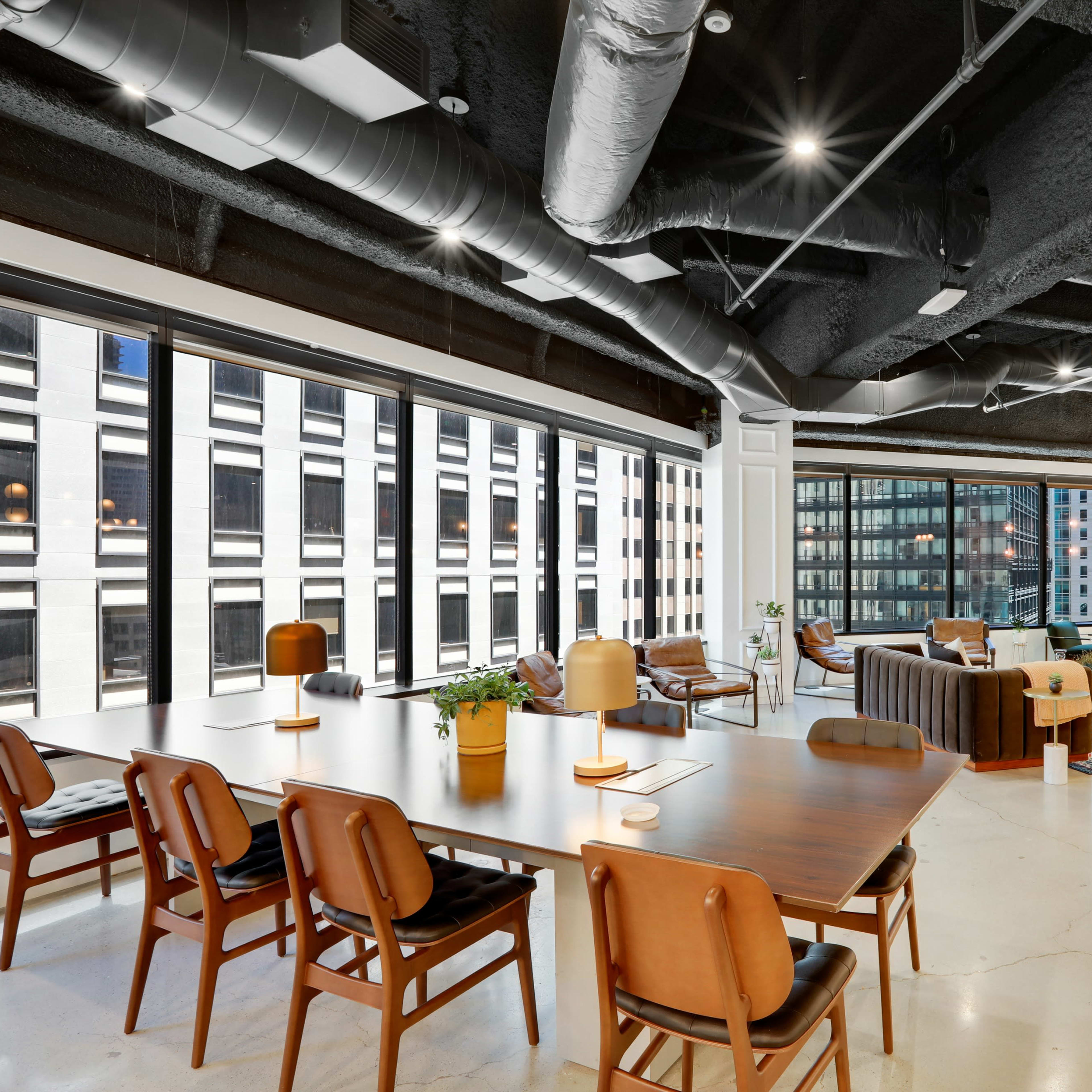 A modern office space featuring a mix of seating areas, large windows with city views, and a dining table surrounded by chairs.