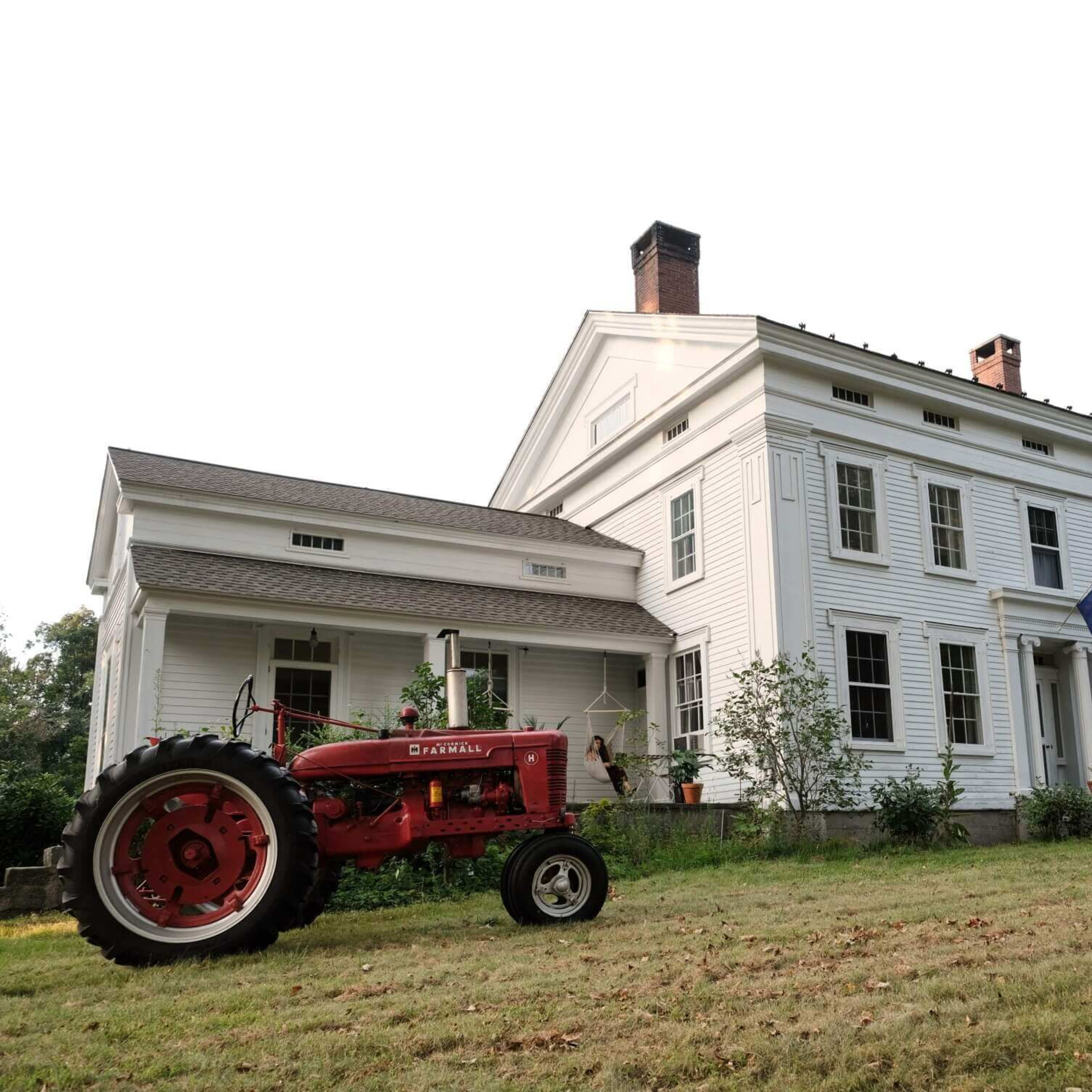 A vintage red tractor rests in front of a large white colonial-style house with a flag displayed.