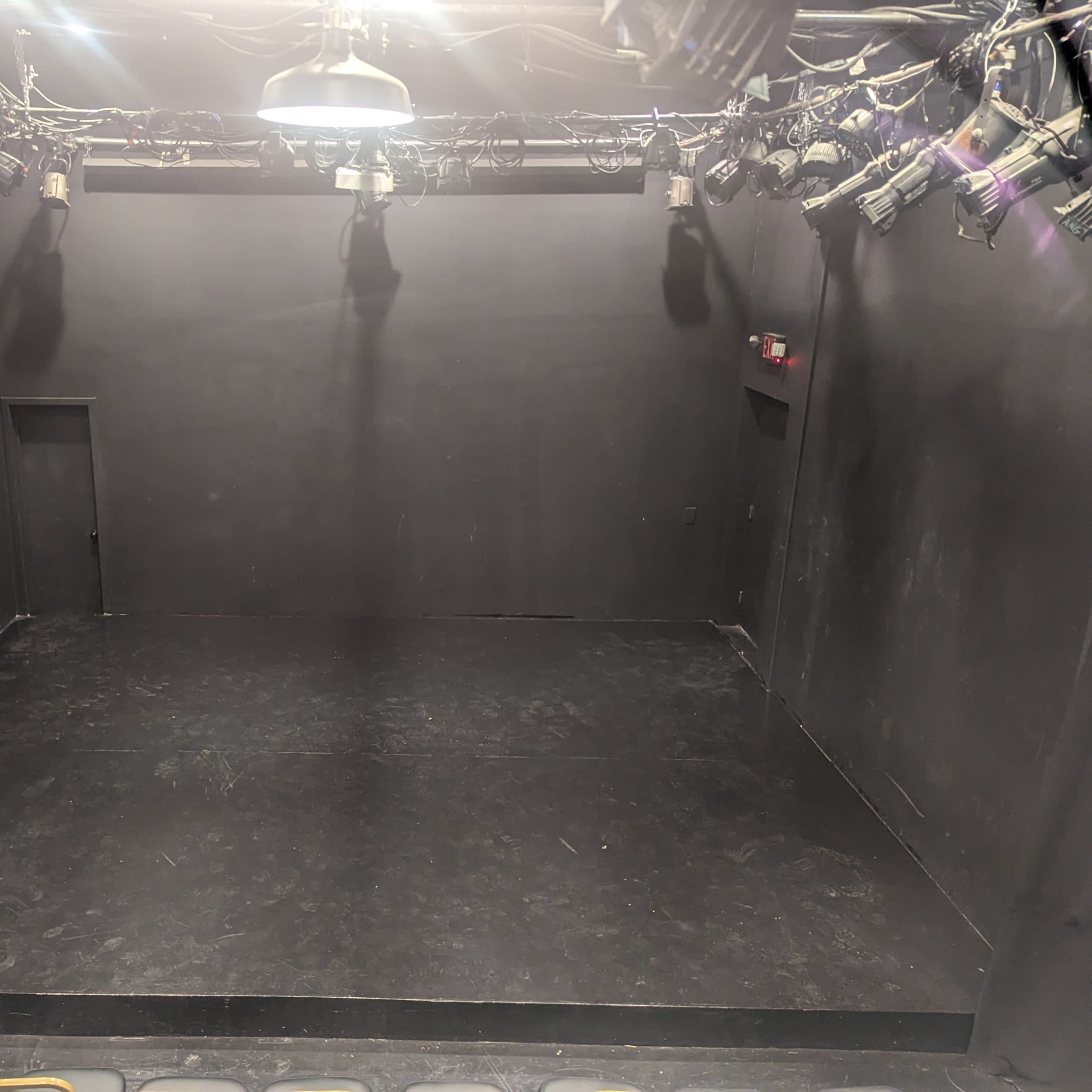 A small, empty black box theater with a few lights hanging from the ceiling and a dark floor.
