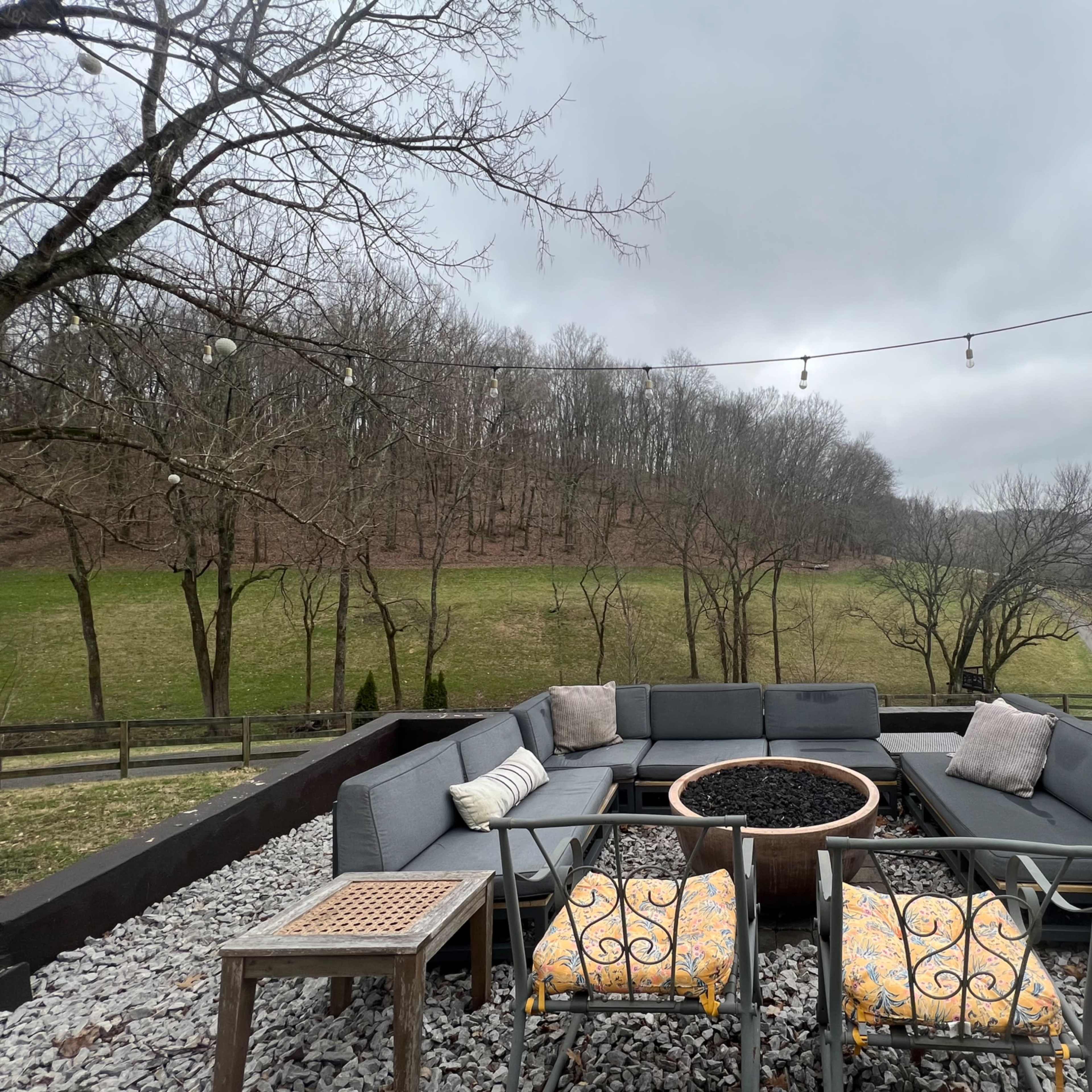 A patio area with grey seating around a circular fire pit, surrounded by gravel and featuring string lights overhead, with a grassy landscape in the background.