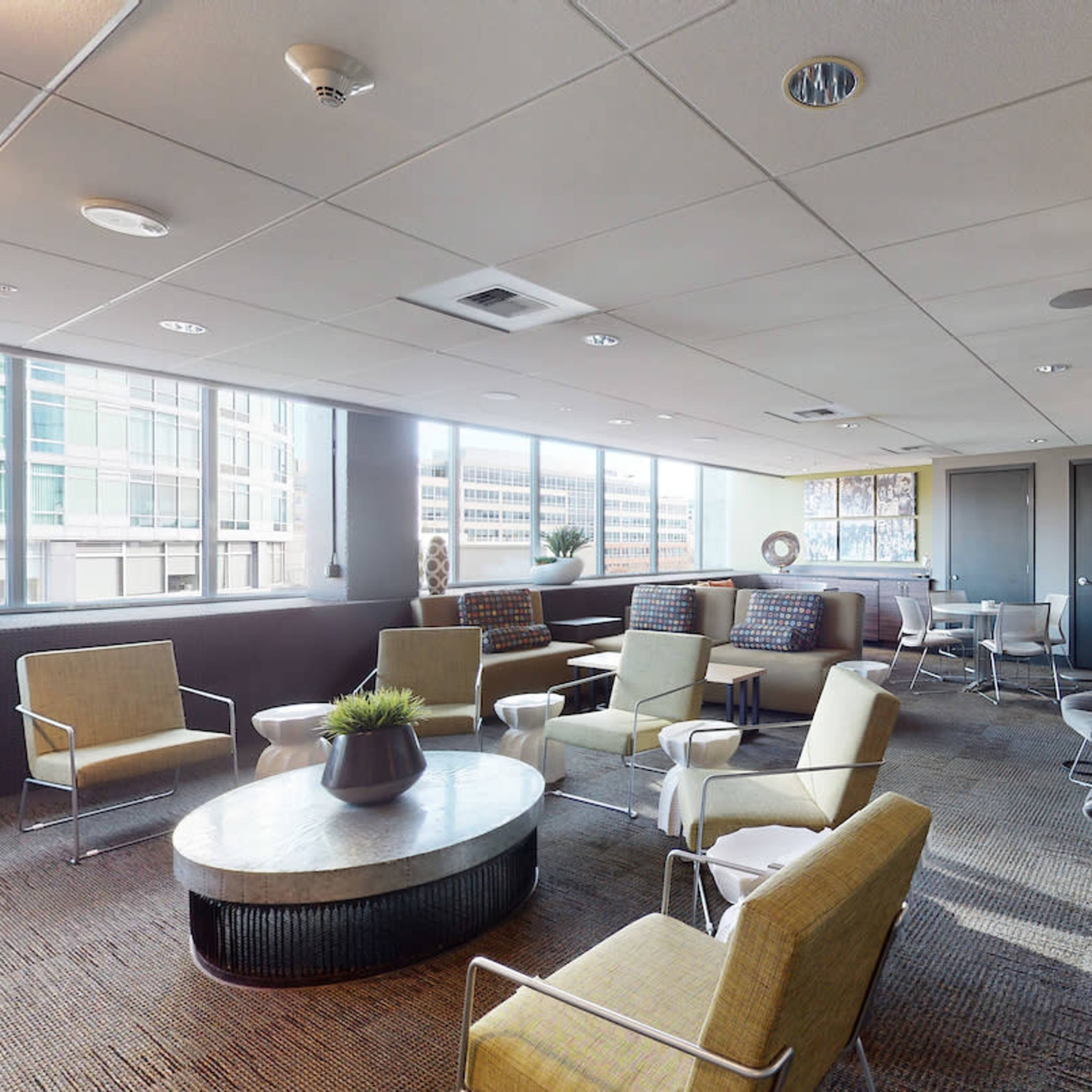 A modern office lounge features multiple seating arrangements with large windows allowing natural light to fill the space.