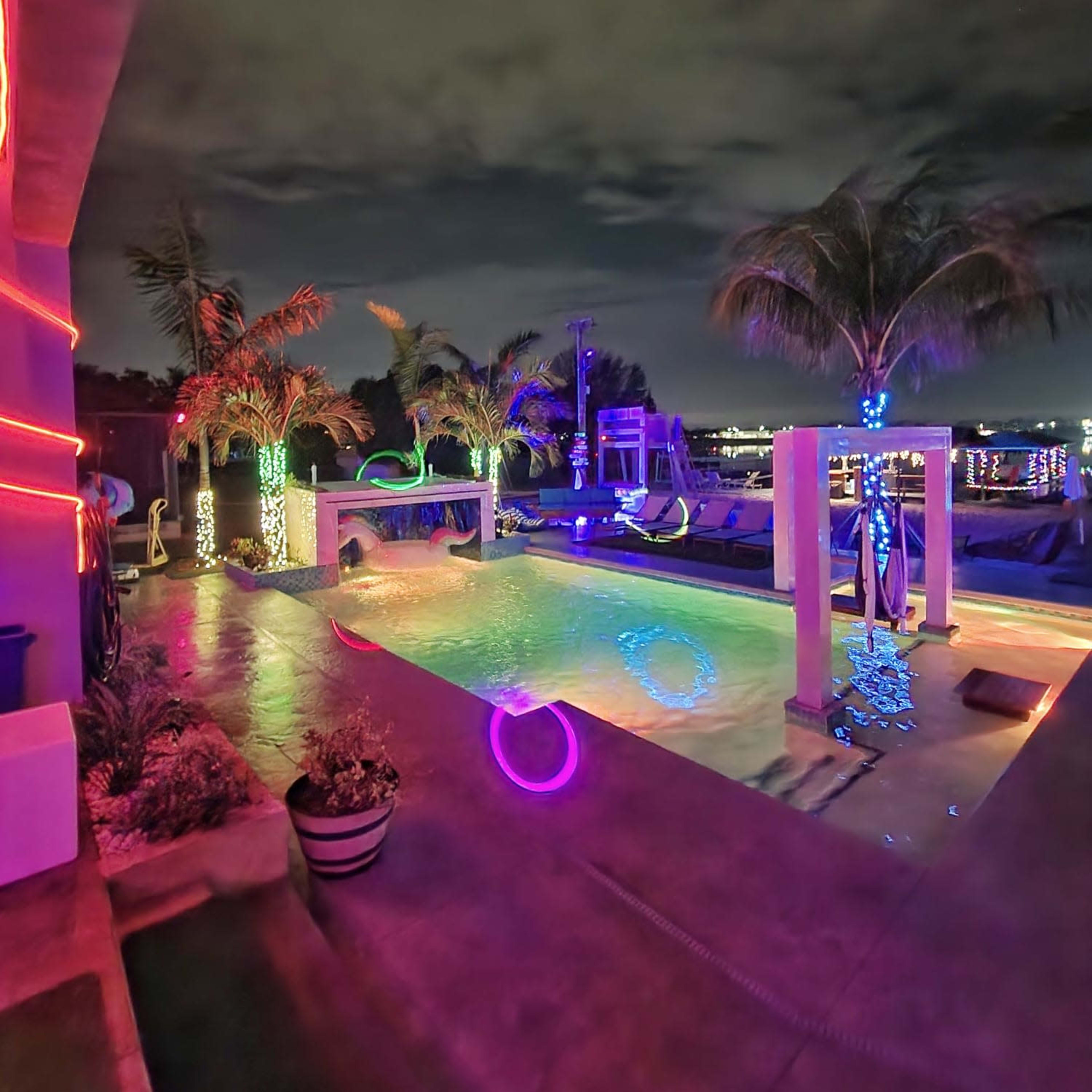 A pool area is illuminated with colorful lights and surrounded by palm trees, set against a night sky.